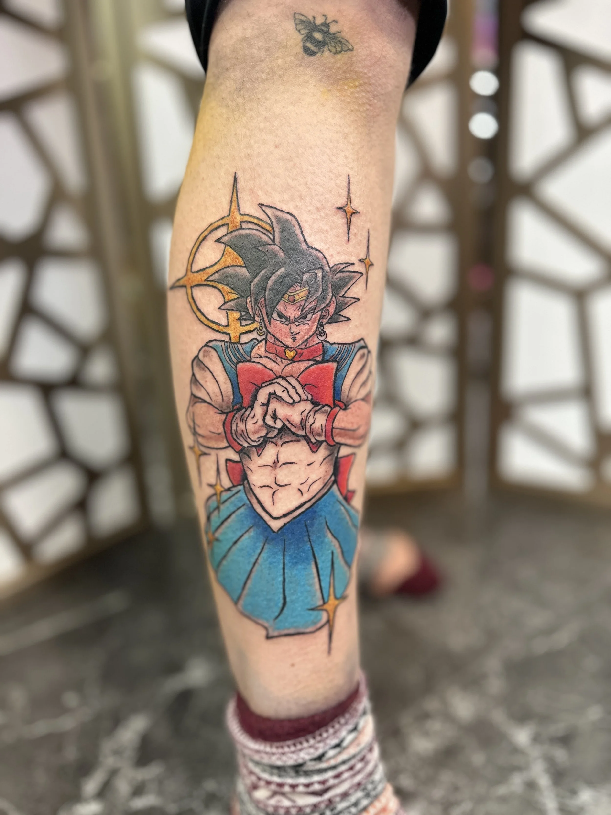 Tattoo of Goku from Dragon Ball Z with a serious expression, standing with arms crossed, wearing Sailor Moon iconic orange and blue outfit, with a halo and star-like sparkles around him.