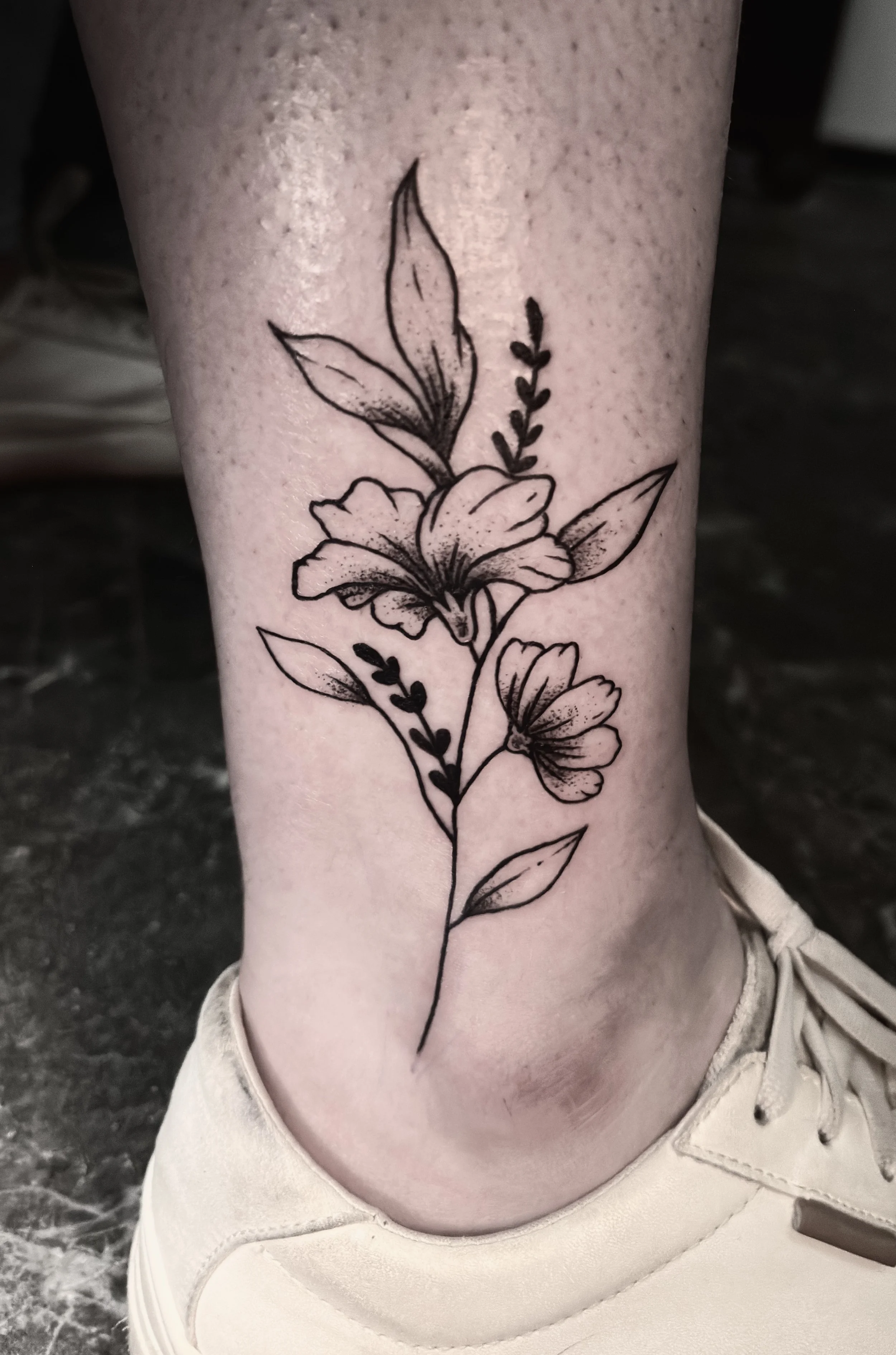 Line art tattoo of a floral arrangement with leaves and flowers on a person's ankle.