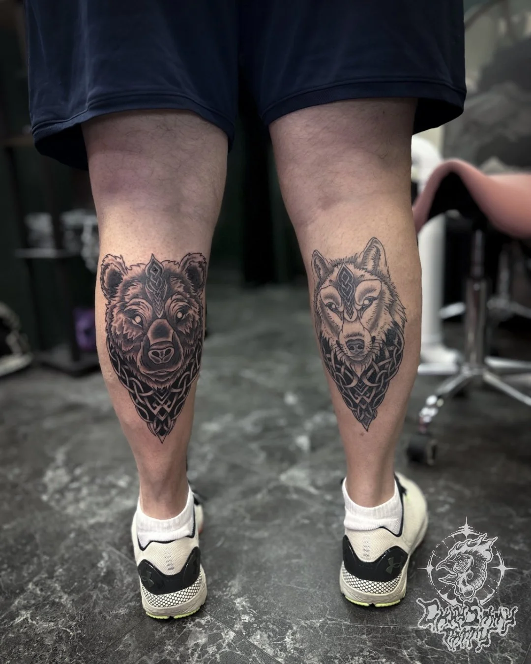 Tattooed bear and wolf portraits on person’s calves, with intricate black line designs, in a tattoo studio.