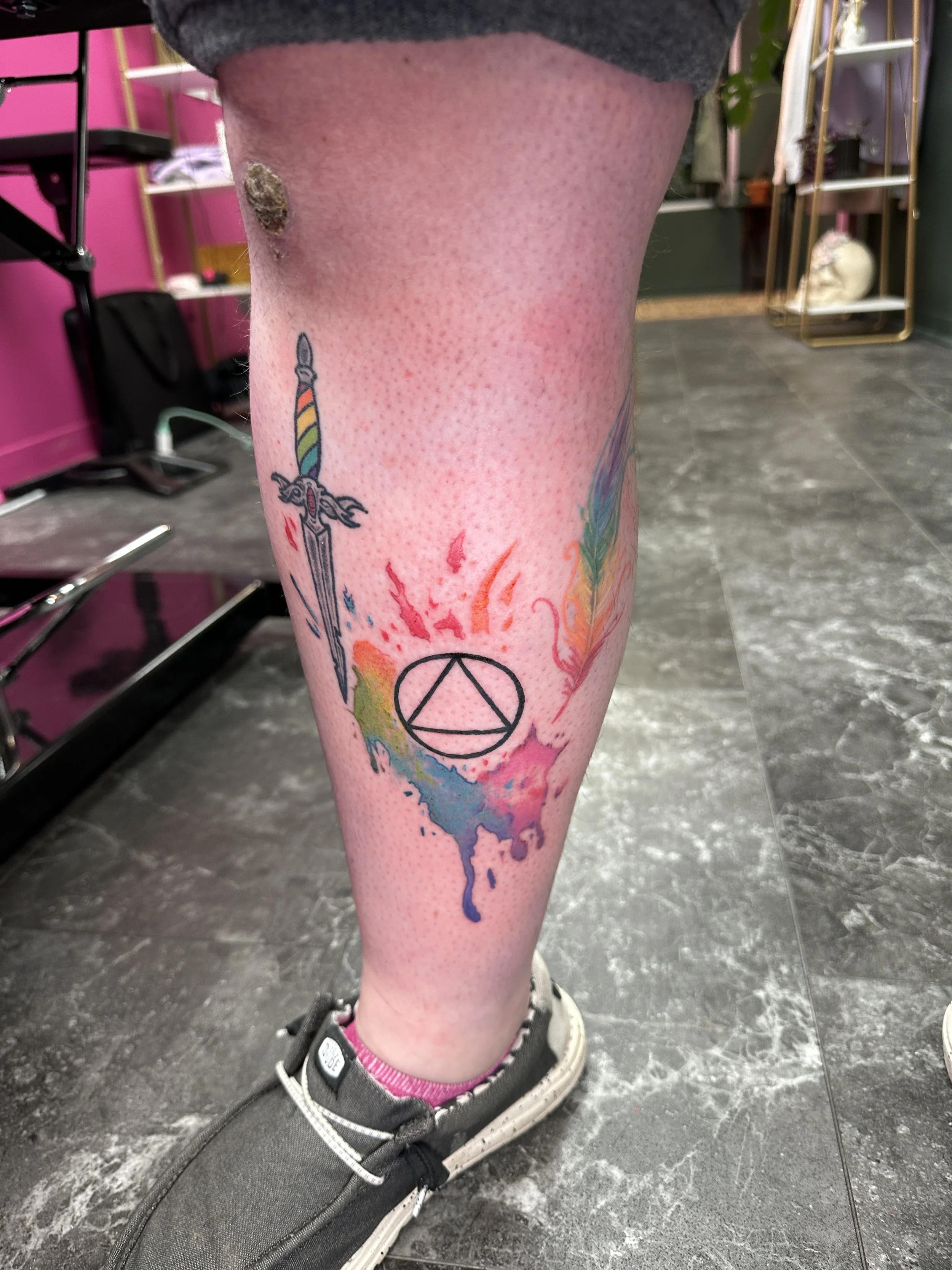 Colorful tattoo featuring a sword with a rainbow-colored hilt, a circle with a triangle inside, and rainbow watercolor splashes on the person's calf, with the person wearing grey sneakers with pink socks.
