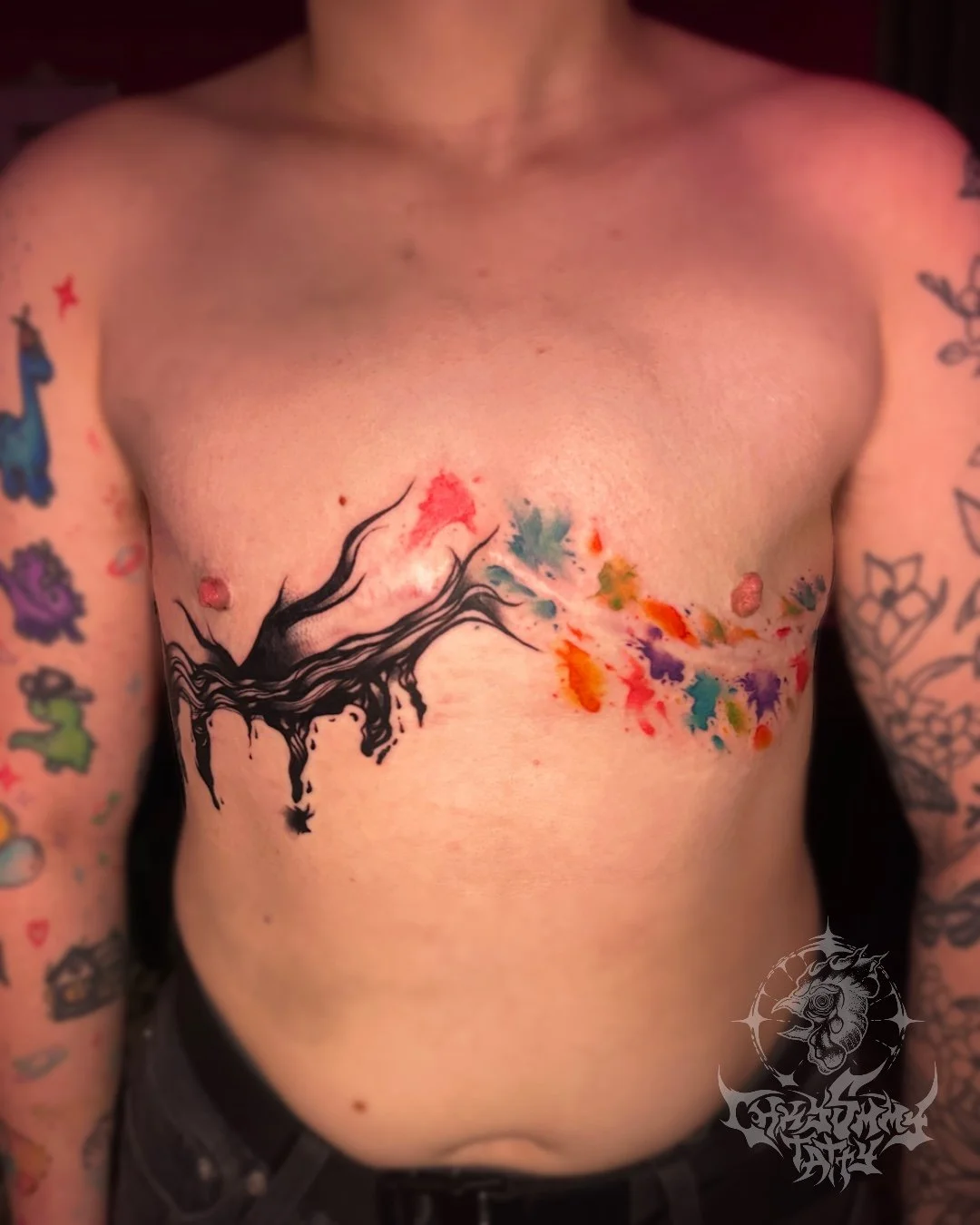 A person's chest with a colorful tattoo of a tree with black branches and multicolored watercolor splashes, with tattoos on both arms.