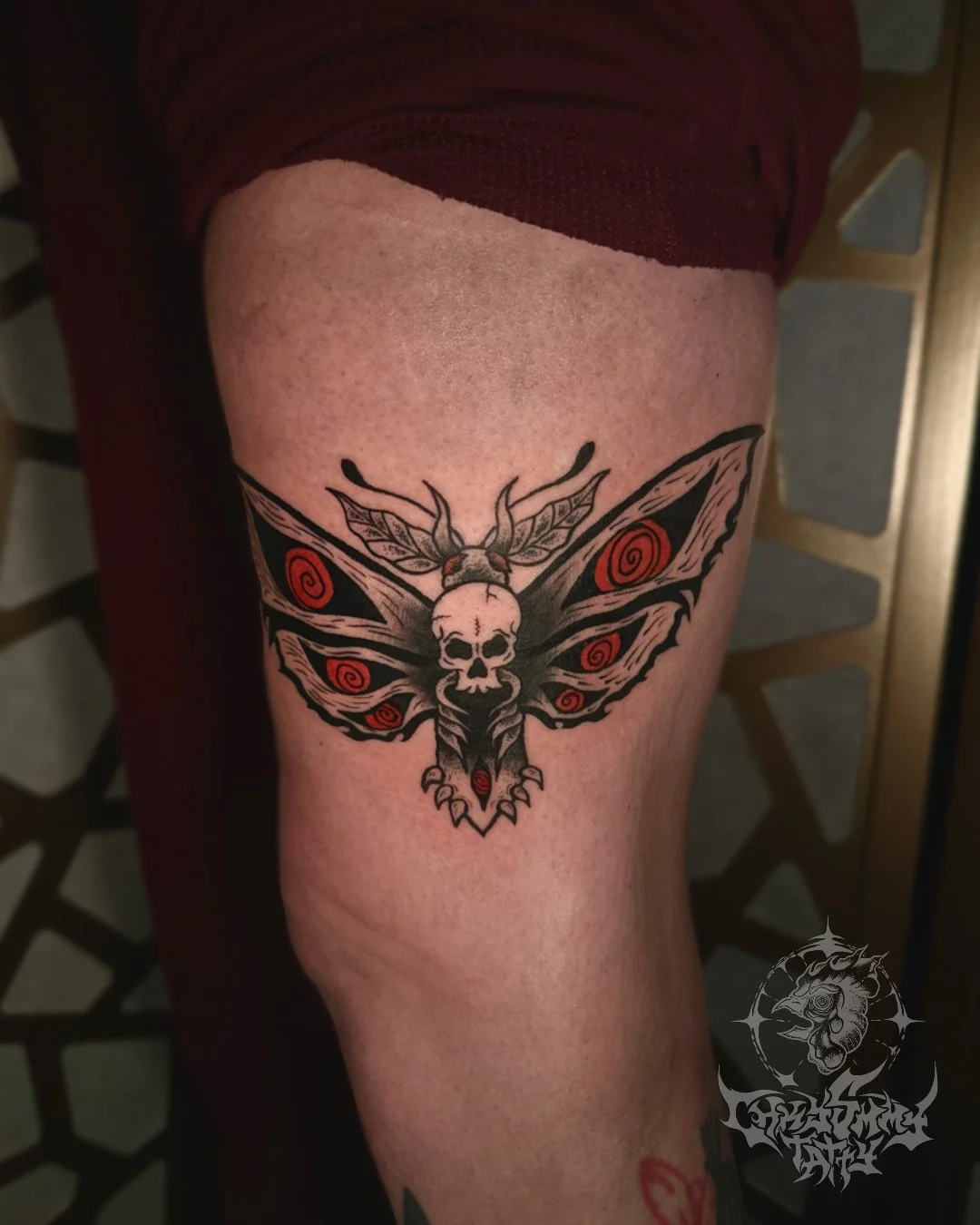 Tattoo of a moth with a skull on its thorax, featuring red spirals on its wings, black and gray shading, on a person's thigh.