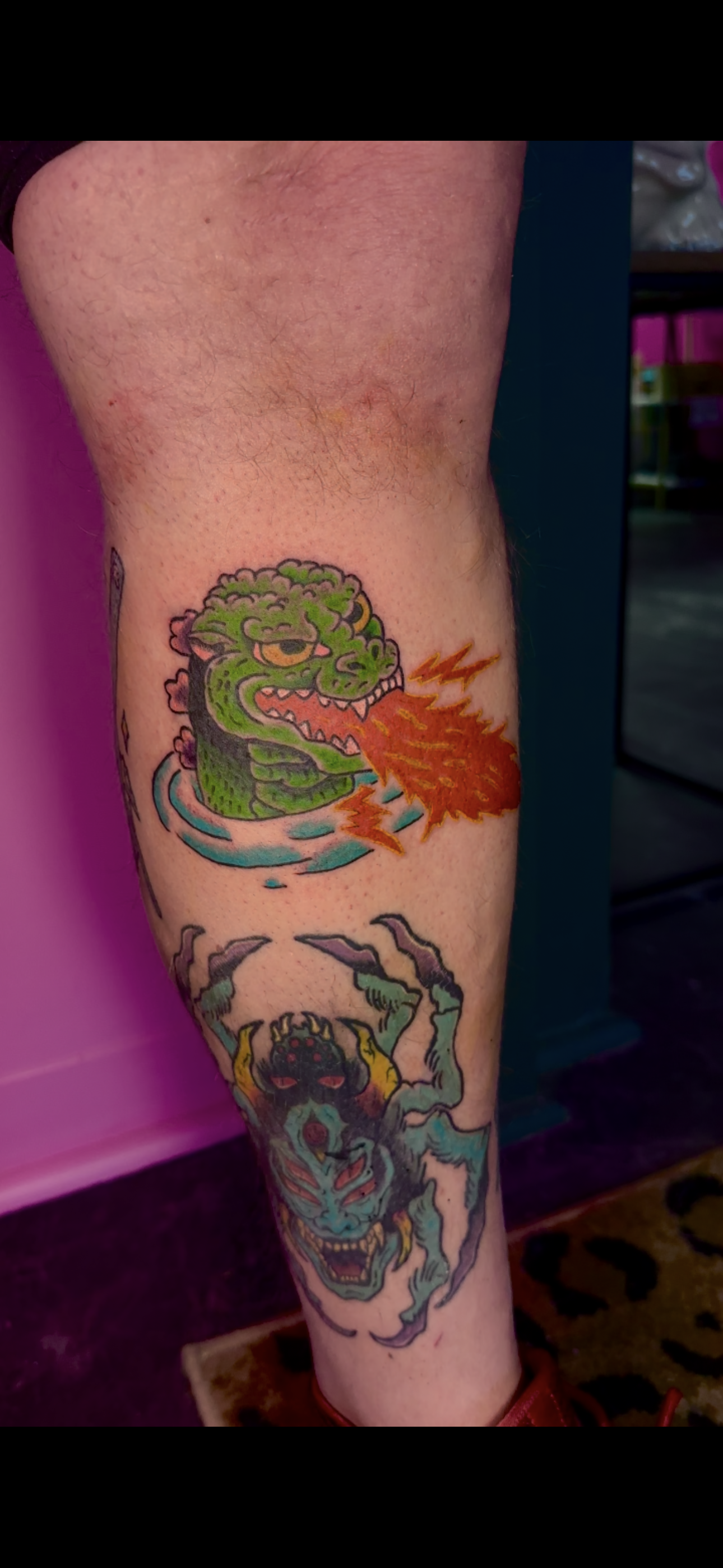 Tattoo of a green cartoonish dragon breathing fire on a person with a blue face, red eyes, and a black demonic appearance. Godzilla American traditional. 