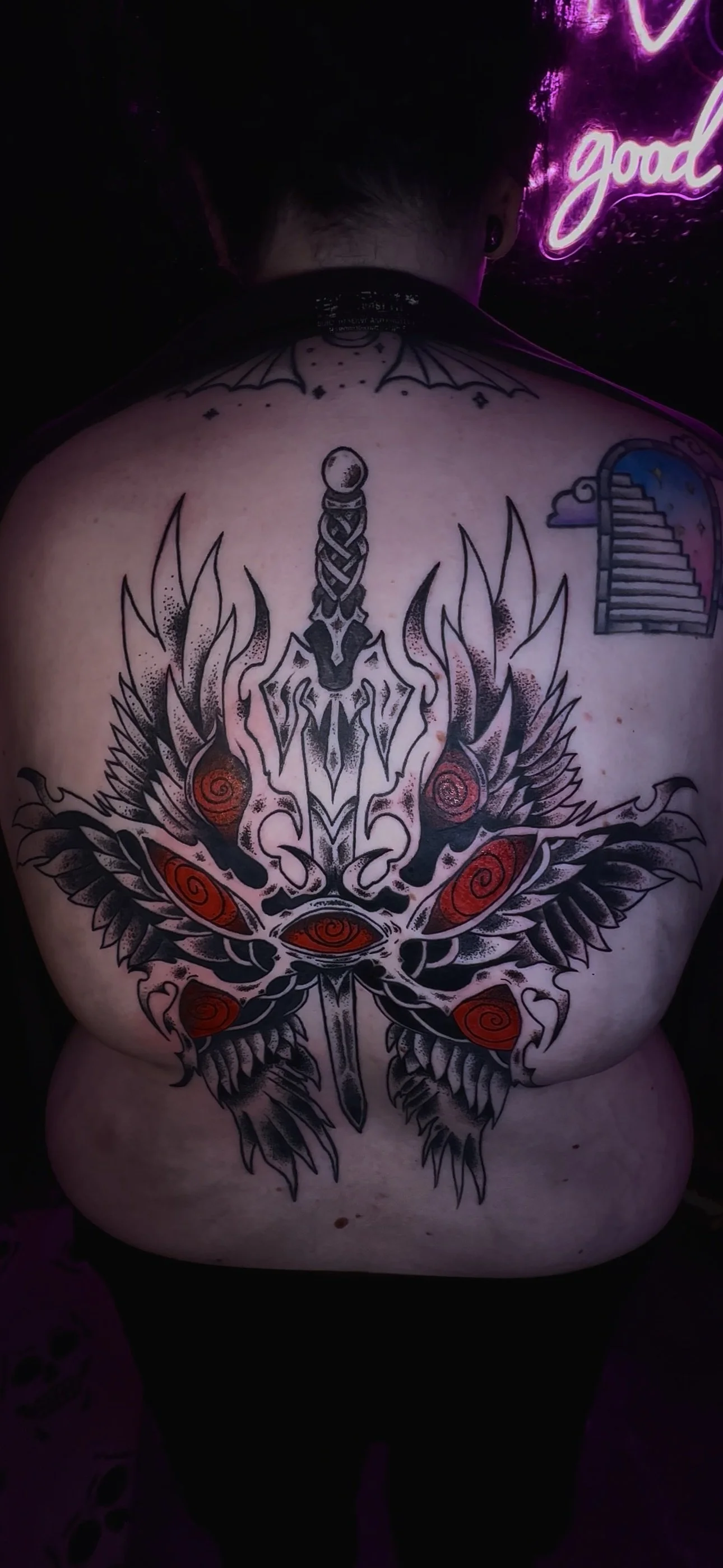 Back tattoo of a symmetrical, black and red design resembling a spider with multiple spiraled eyes and intricate details, with a neon purple light and a staircase illustration in the background.
