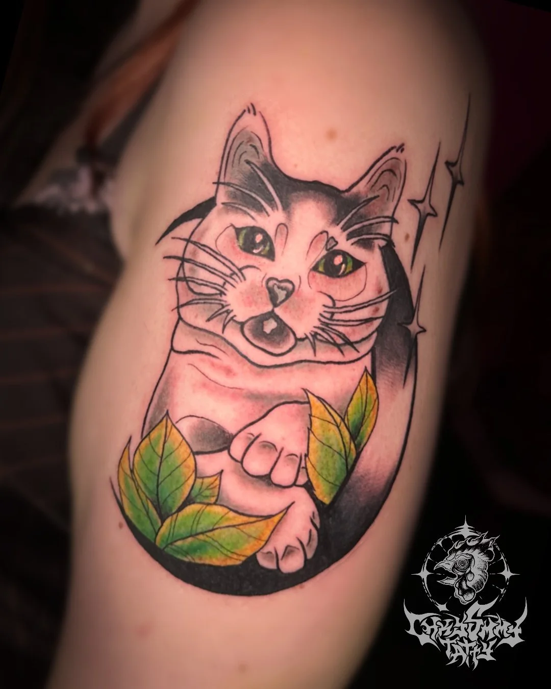 Colorful tattoo of a playful cat with green eyes, black and pink fur, surrounded by yellow and green leaves, and sparkles.