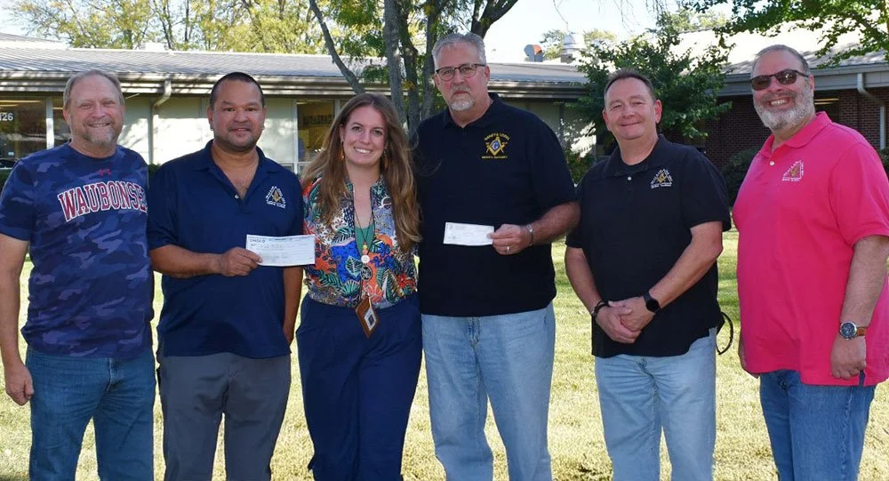 Six people standing outside on a grassy area, posing for a photo. The group consists of five men and one woman, with some holding checks. They are smiling are celebrating a donation. There are trees and a building in the background.