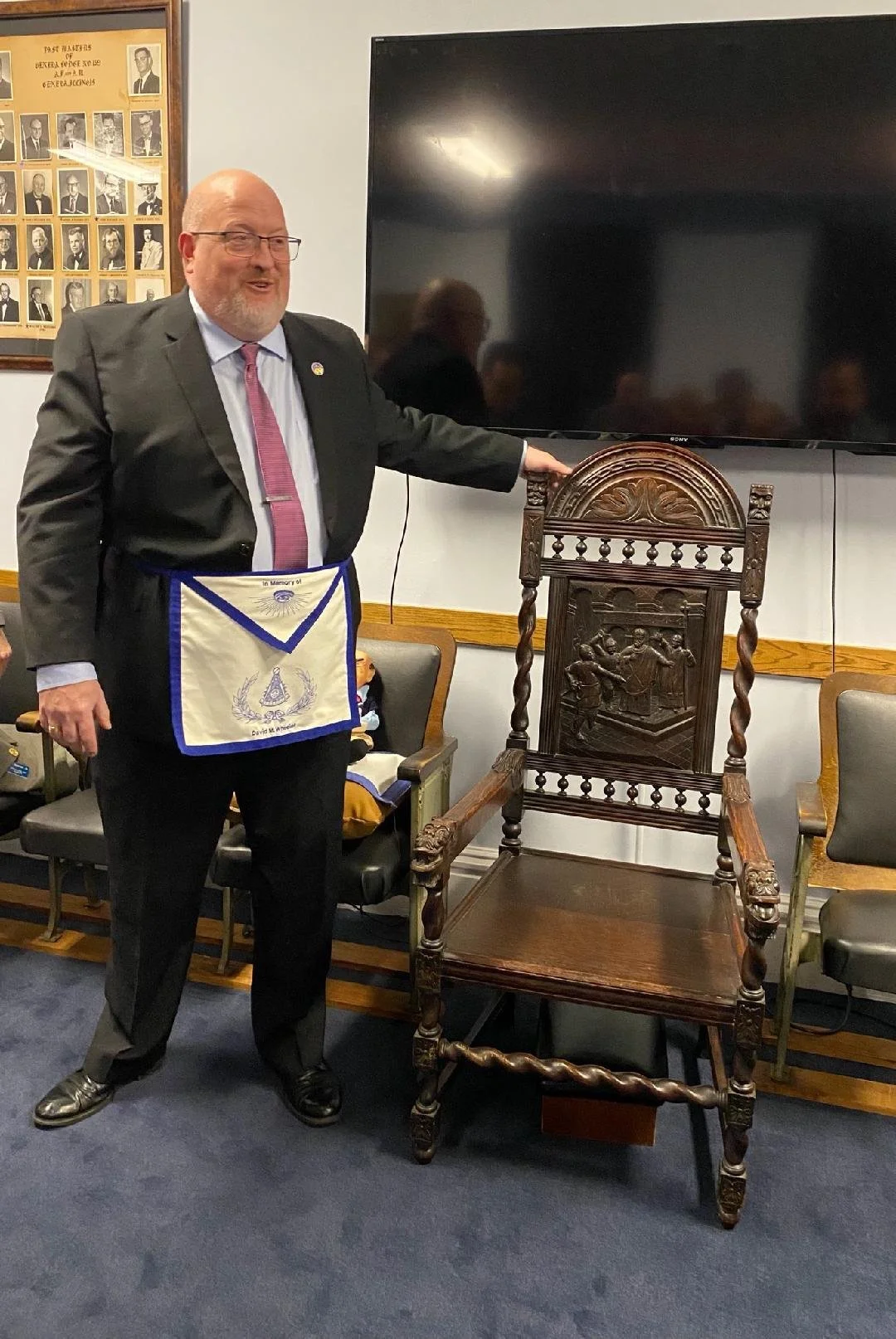 A man in a suit and pink tie standing next to an ornate wooden chair, touching the top of it, in a room with a framed photo collage on the wall and a TV screen.