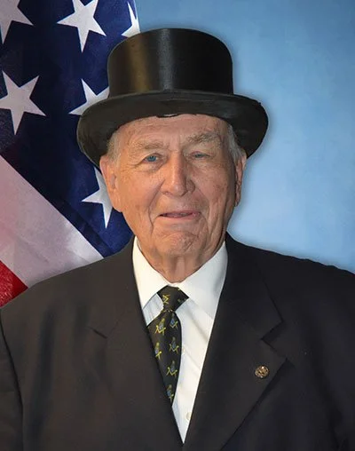 An elderly man dressed in a formal suit and tie, wearing a black top hat, standing in front of a United States flag, with a blue background.