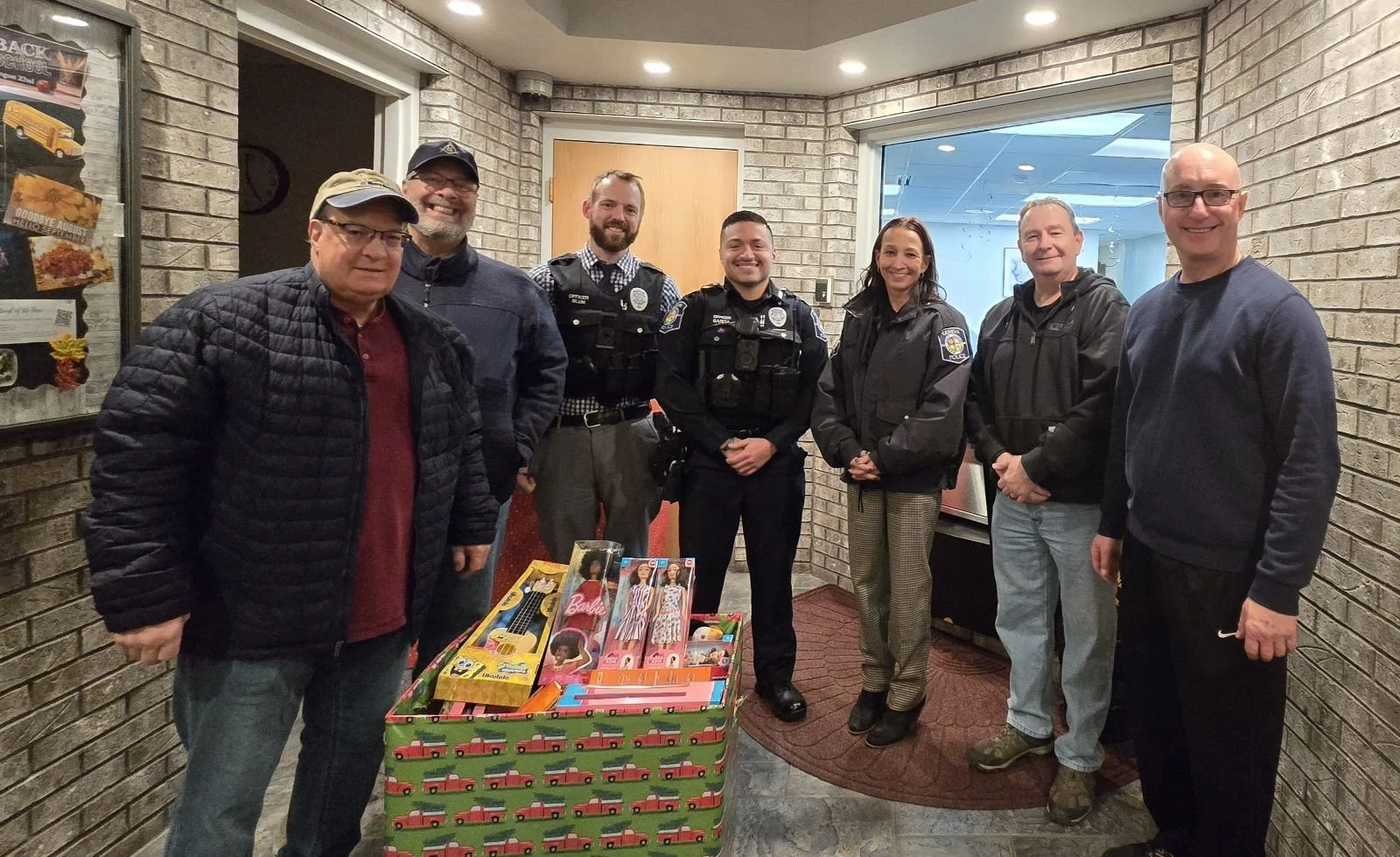 Group of eight people, including police officers and civilians, standing inside a building, smiling, with a large wrapped box of toys in front of them.