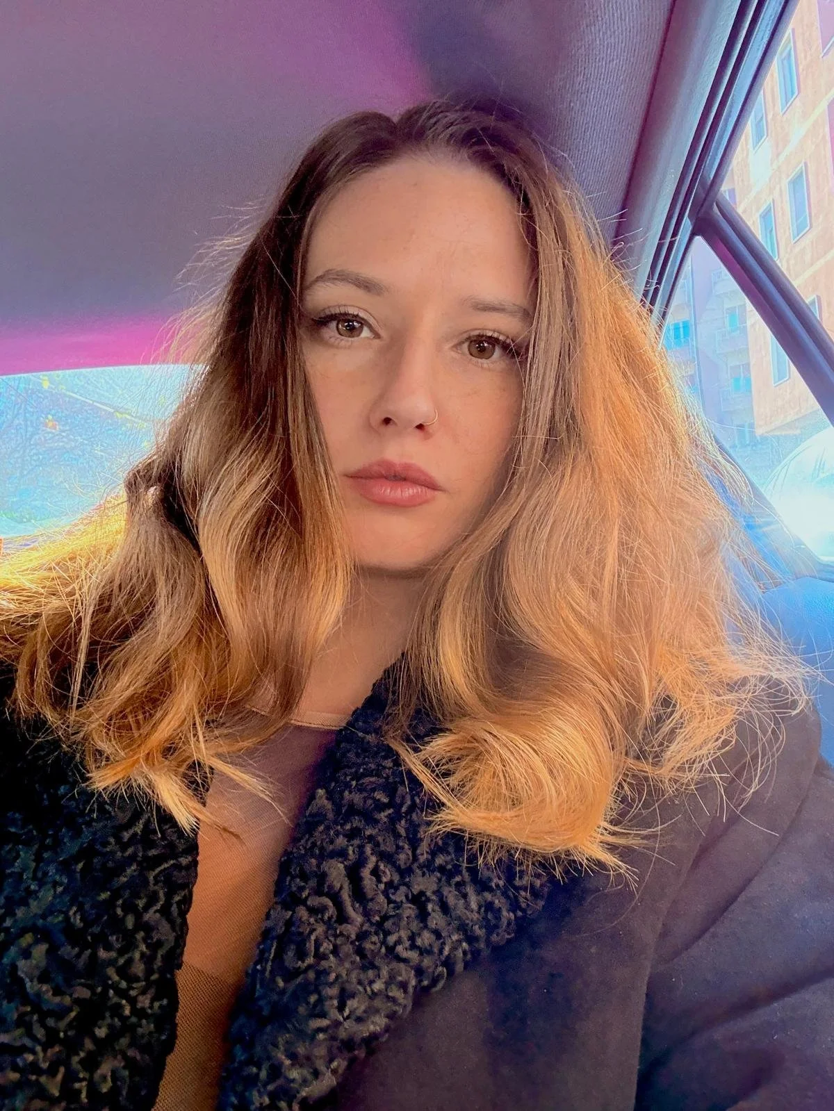 Retrato de una mujer con cabello rubio y rizado dentro de un coche, con una ventana mostrando edificios urbanos al fondo y una luz suave iluminando su cara.