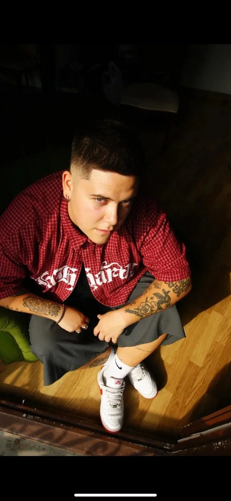 Joven con cabello corto y tatuajes en los brazos, vistiendo una camisa roja con cuadros y pantalones cortos grises, sentado en el suelo de madera y con una mirada intensa.