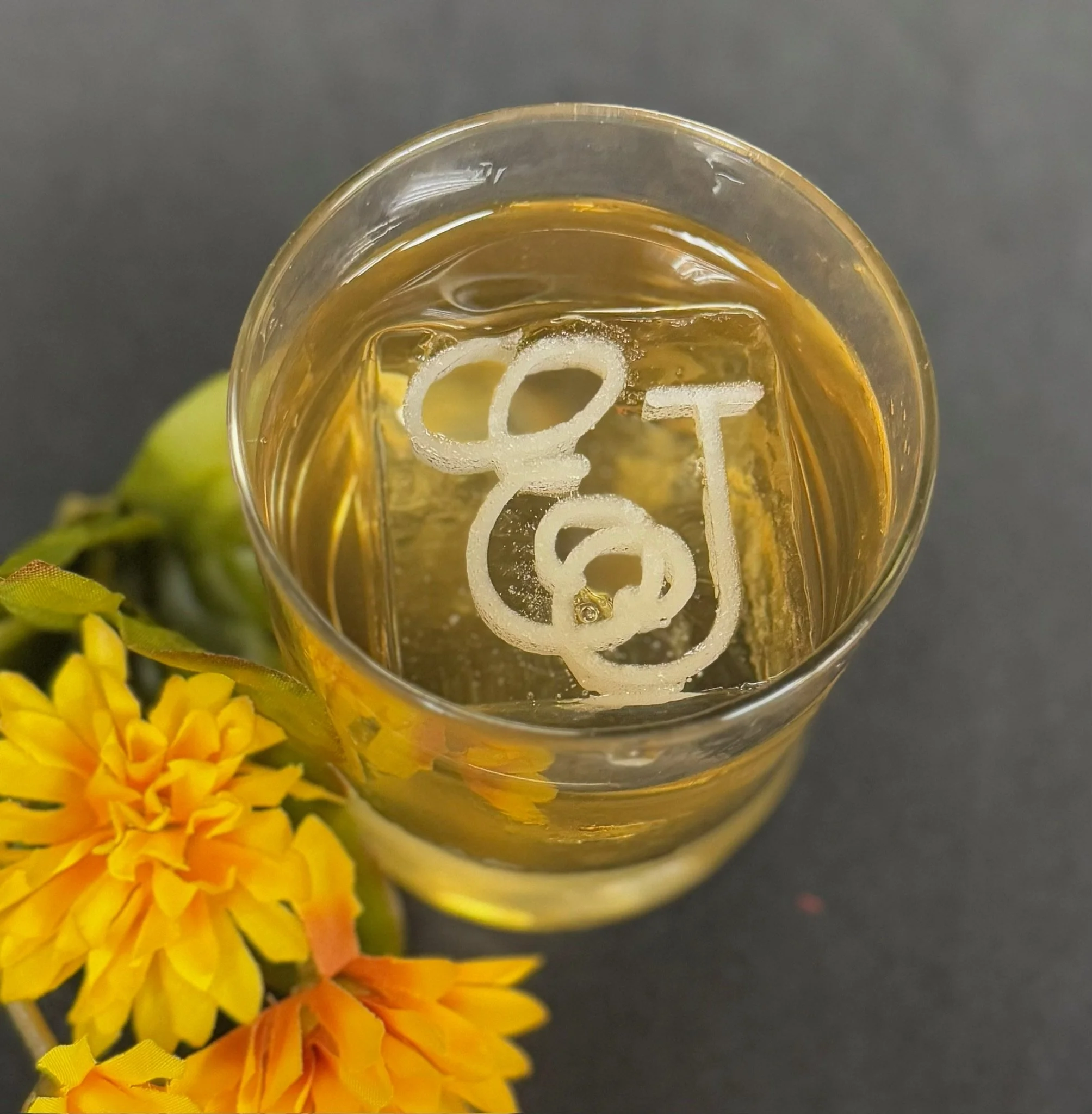 Close-up of a glass with a yellow beverage, custom ice cubes, and lemon slices, with yellow flowers in the background.