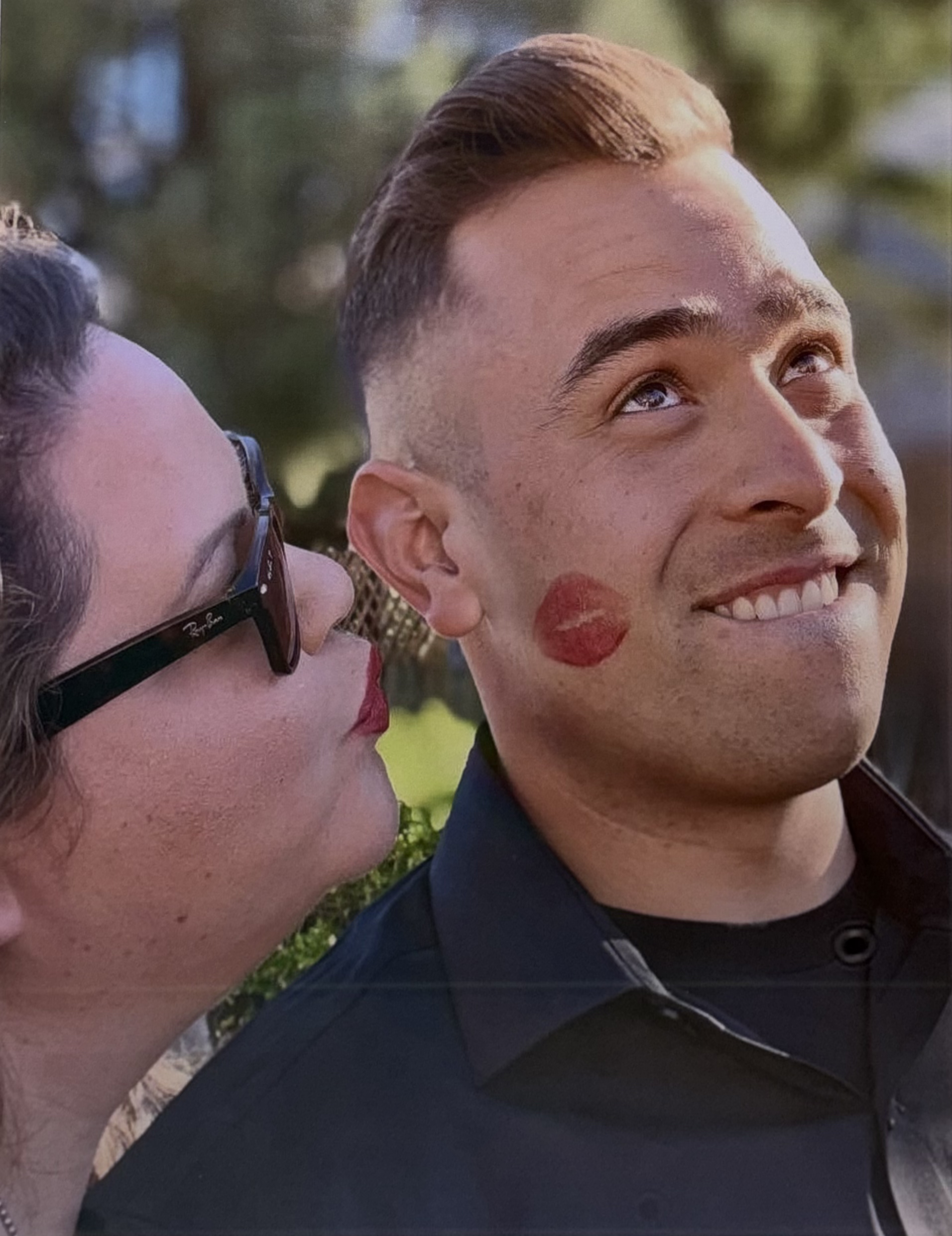 A woman with curly hair, glasses, and lipstick looks at a smiling man with a serious expression who has a lipstick kiss mark on his cheek.