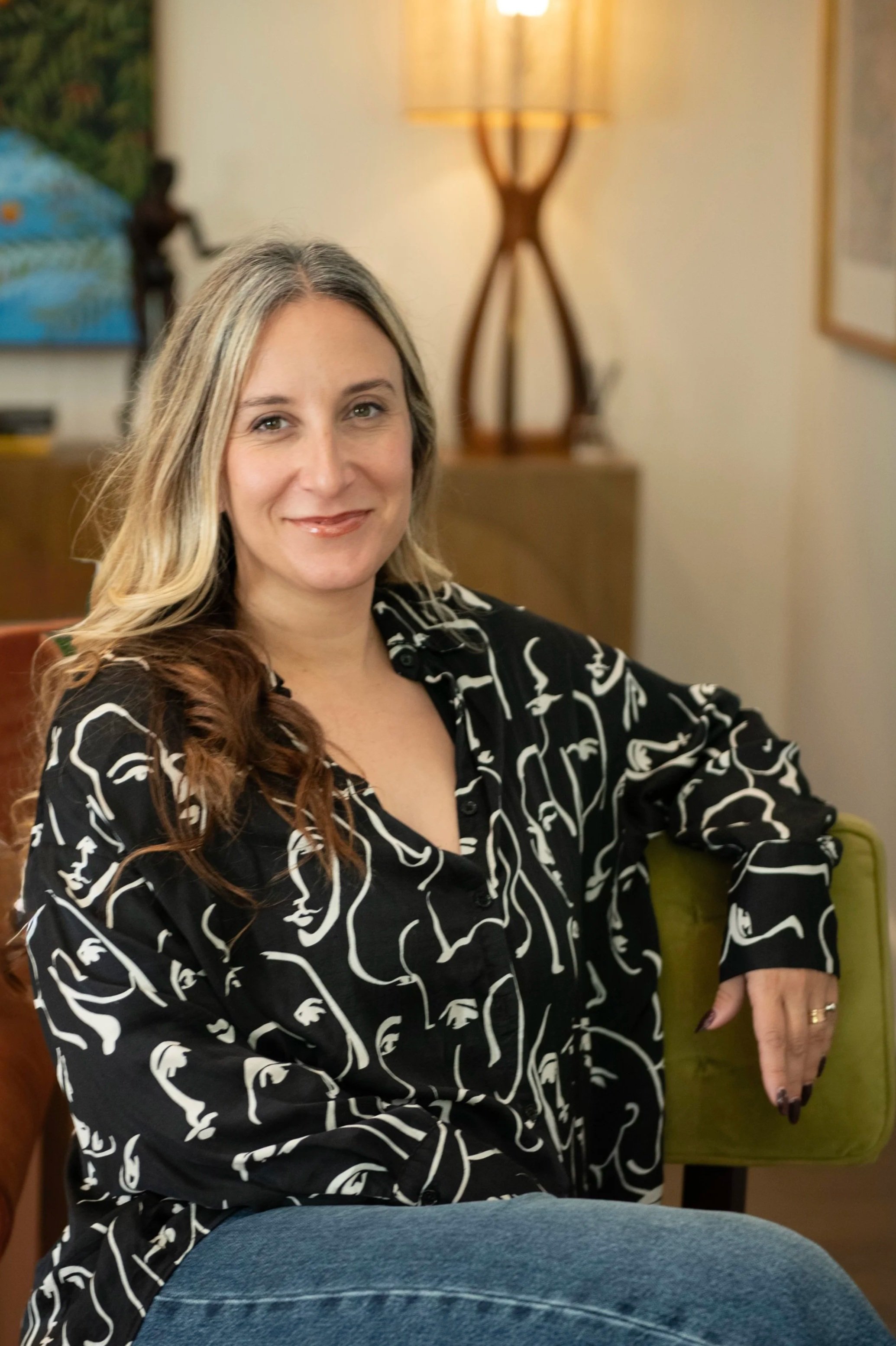 A woman with long, wavy hair sitting on a green chair, smiling, in a room with a beige wall, a lamp, and a painting in the background.