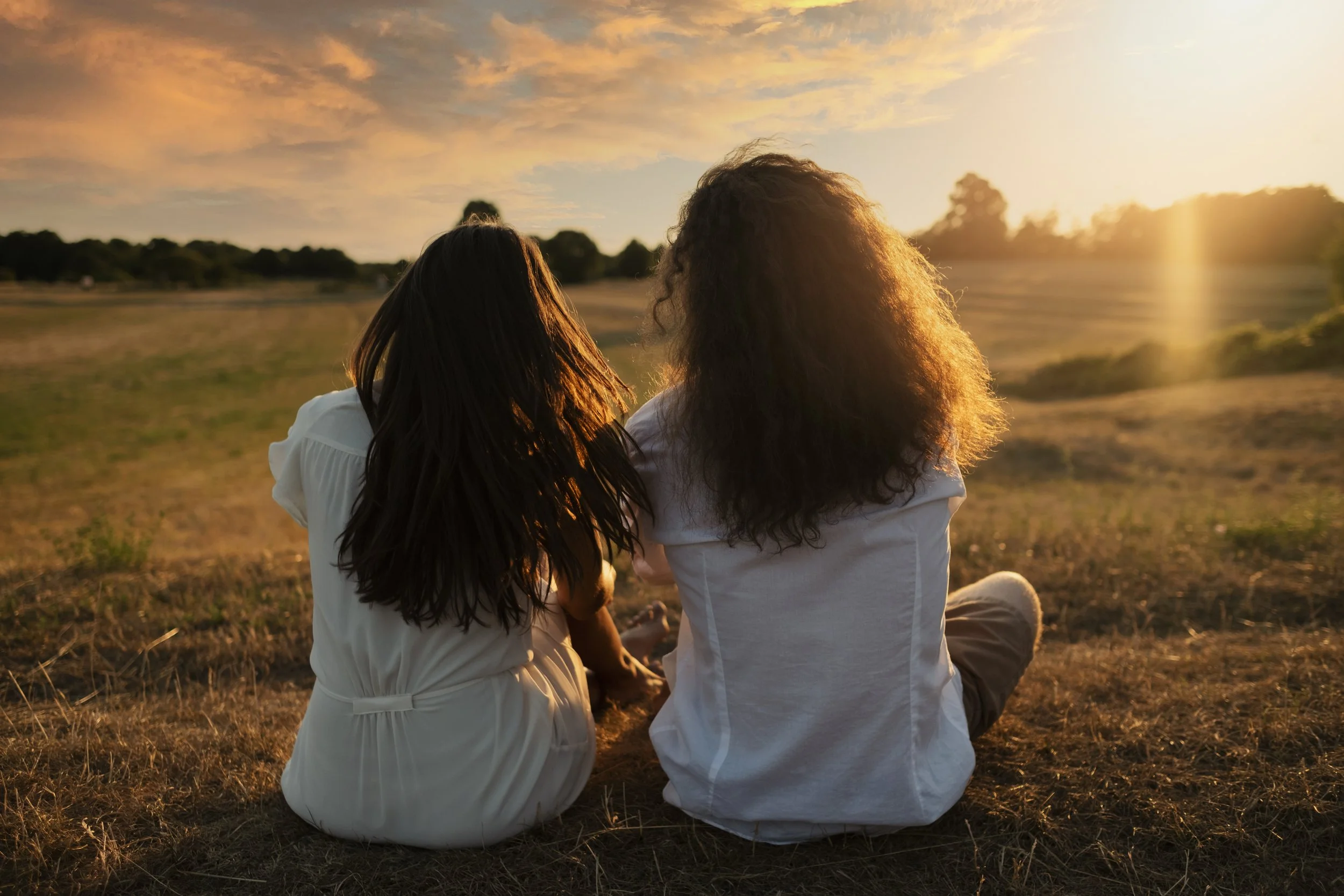 Two women with long hair sitting on the grass during sunset, facing away from the camera, in a field with a partly cloudy sky.