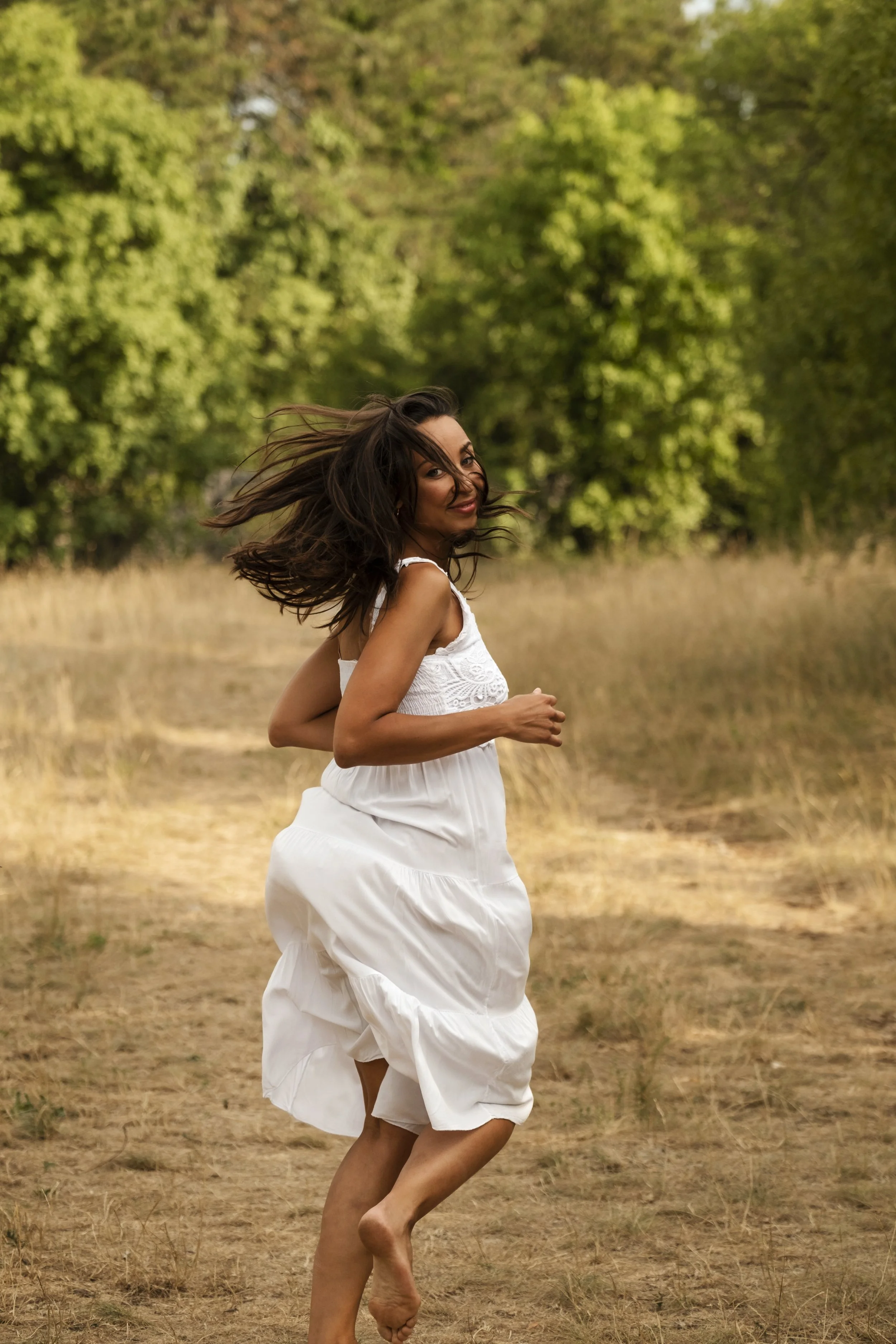 A woman with long dark hair running outdoors in a field, wearing a white dress, with green trees in the background.
