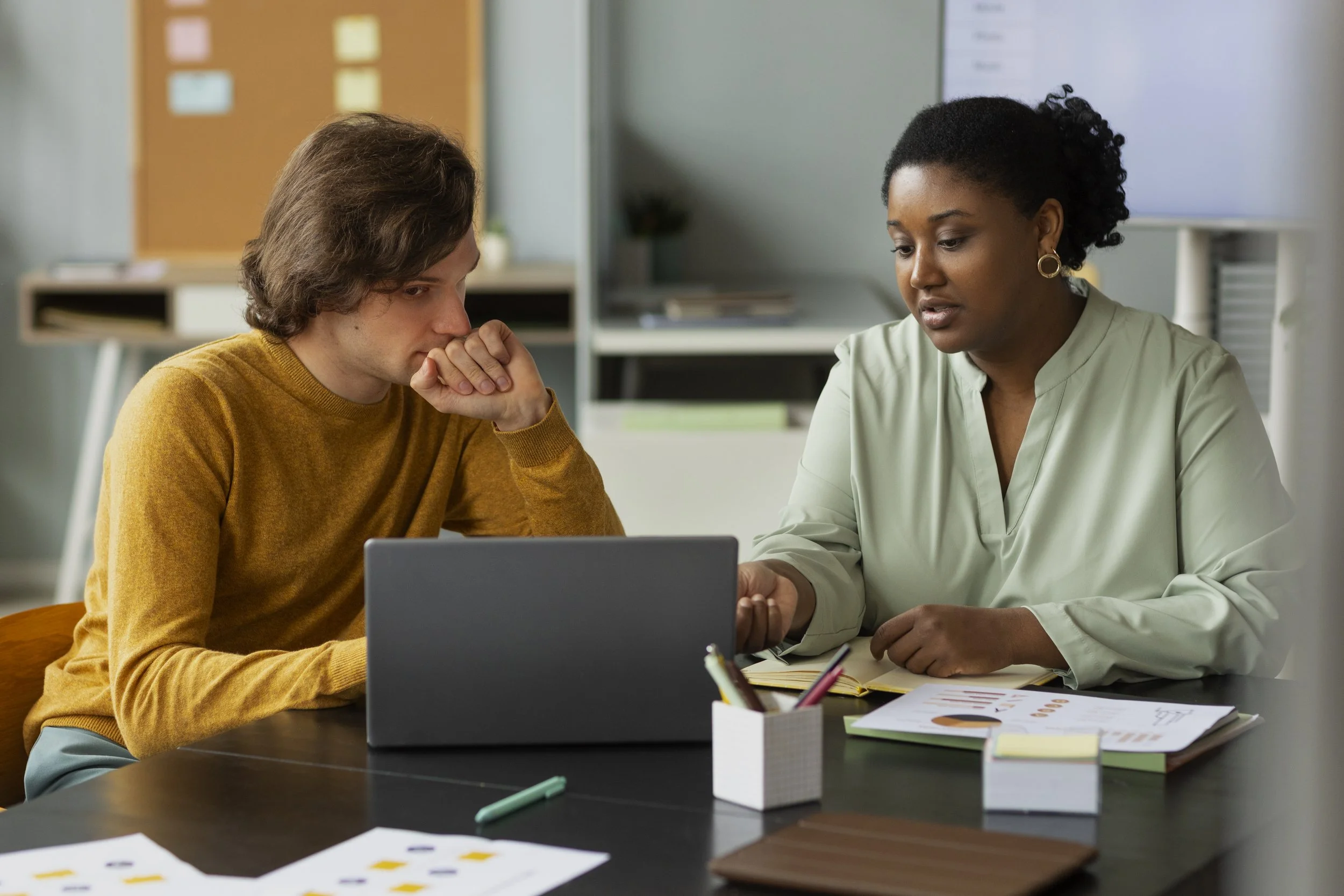A woman and a man are sitting at a desk, looking at a laptop screen, engaged in a discussion in an office setting.