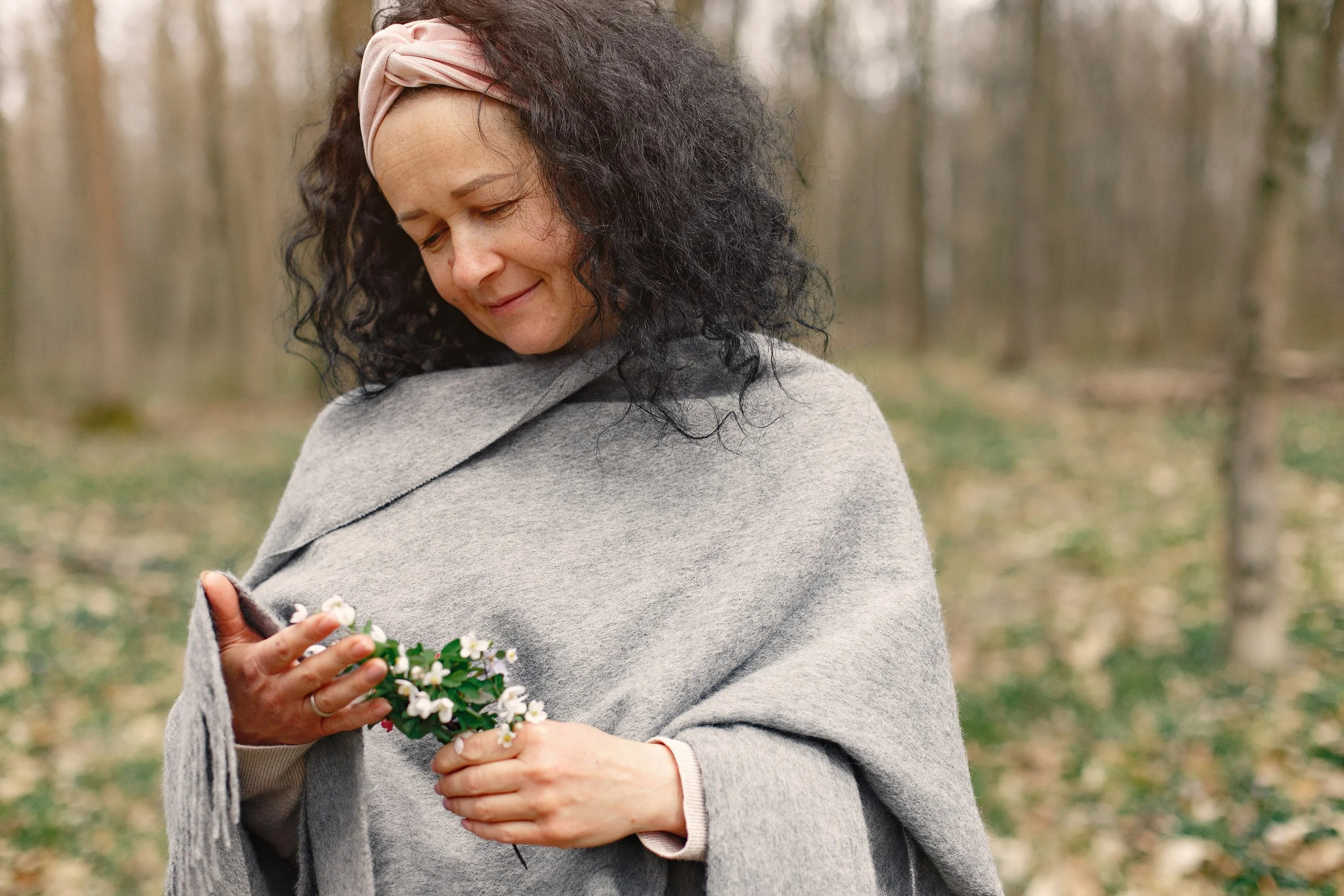 A woman with curly dark hair wearing a pink headband, gray coat, and beige sweater, holding a small bouquet of white flowers in an outdoor wooded area.