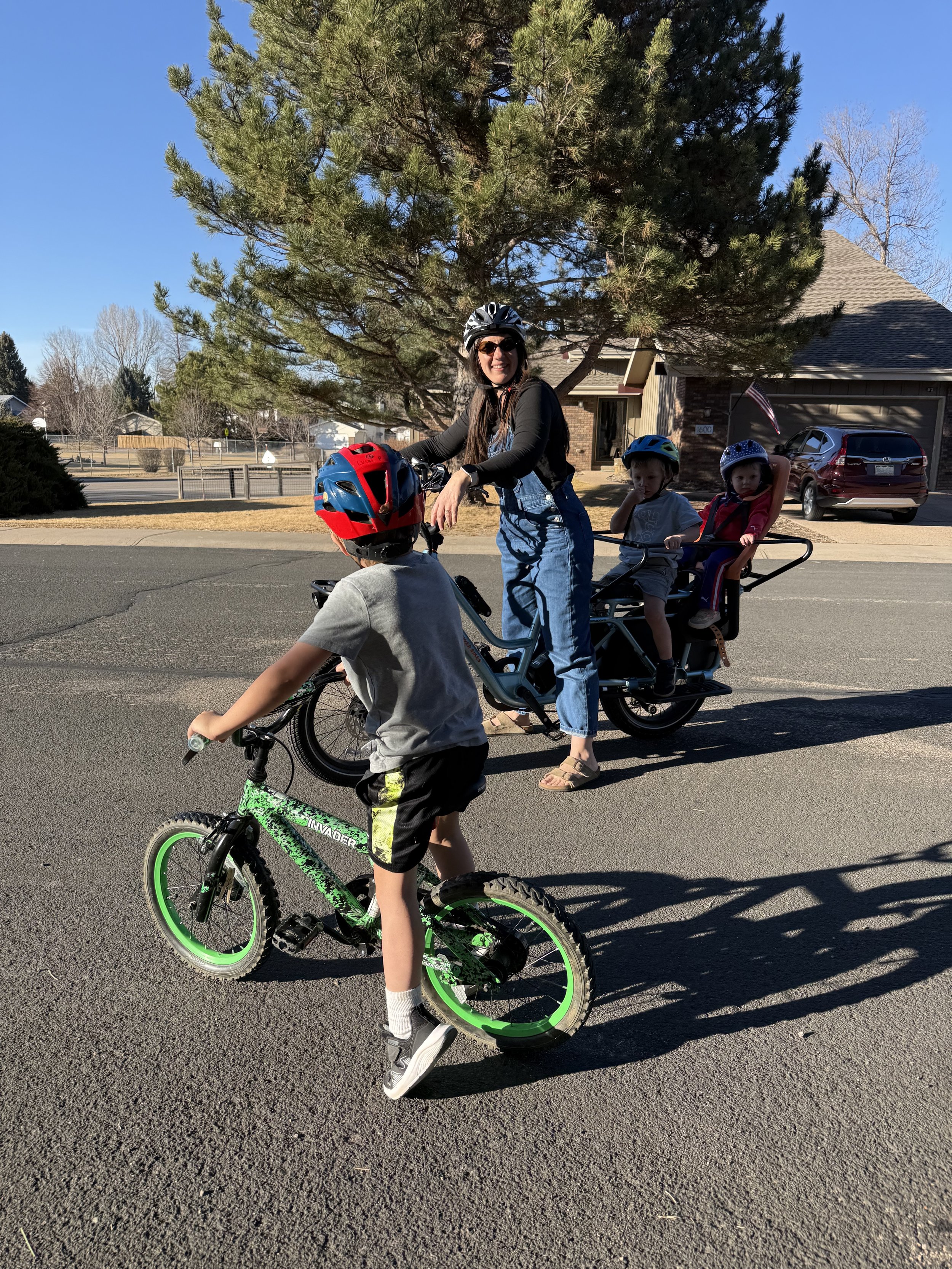 A woman in sunglasses and overalls is smiling while riding a pedal bike with three children, one riding a smaller bike and two seated in a custom pedal stroller, on a suburban street on a sunny day.