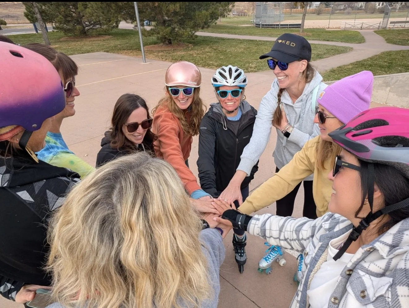 Group of smiling women in helmets and sunglasses placing hands together in a circle outdoors at a park, some with roller skates.