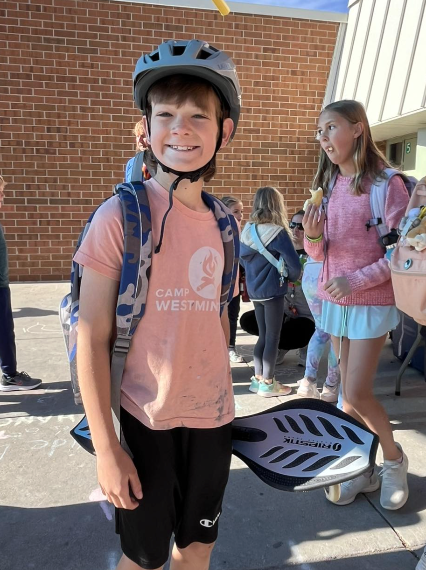 A young boy wearing a bike helmet and a pink Camp Westminister t-shirt, smiling at the camera, standing outdoors with other children in the background.