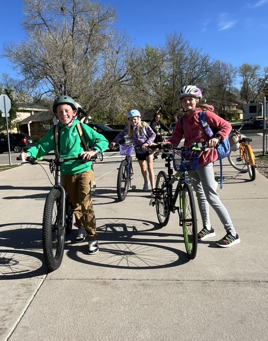 Four children with helmets and backpacks standing with bicycles on a sidewalk, smiling, with trees and parked cars in the background on a sunny day.