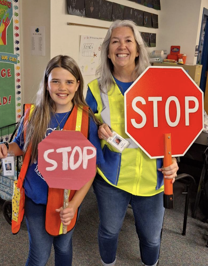 A young girl and an older woman standing inside a classroom, both holding bright red stop signs. The girl has long light brown hair and is wearing a blue shirt and orange safety vest, while the woman has gray hair and is wearing a safety vest over a blue shirt. They are smiling at the camera.