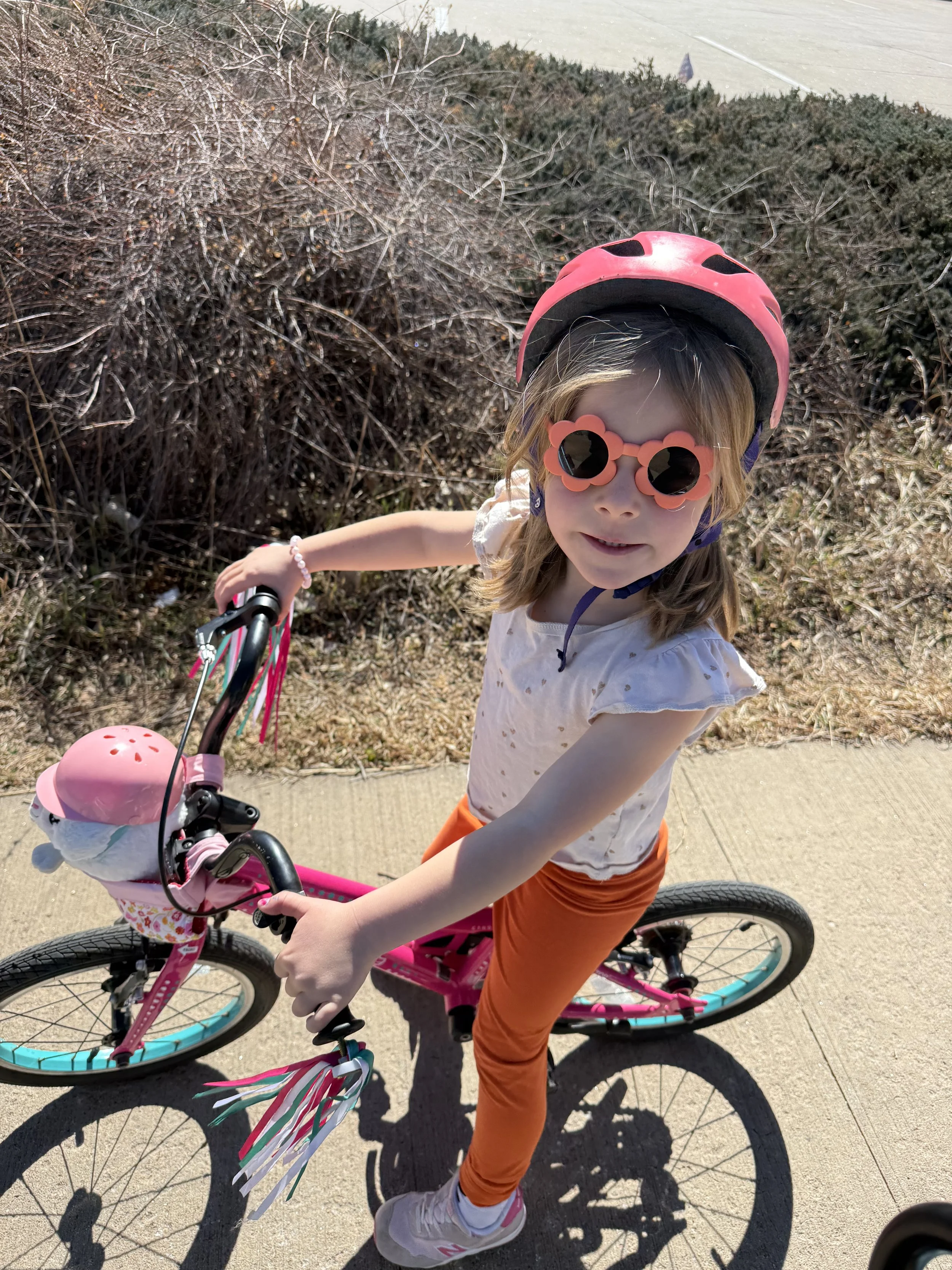 A young girl wearing a pink helmet and flower-shaped sunglasses riding a pink bicycle on a sidewalk.