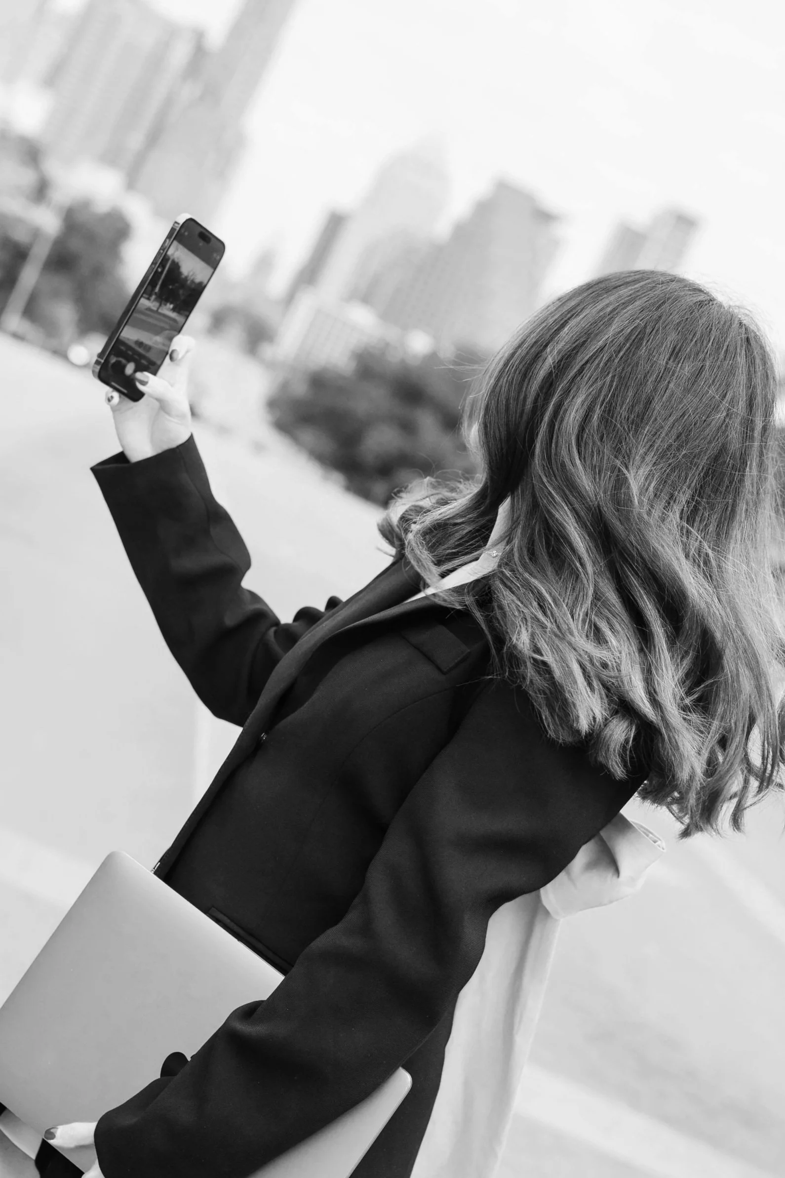 A woman with wavy hair holding a smartphone taking a selfie or photo outdoors in an urban setting in black and white.
