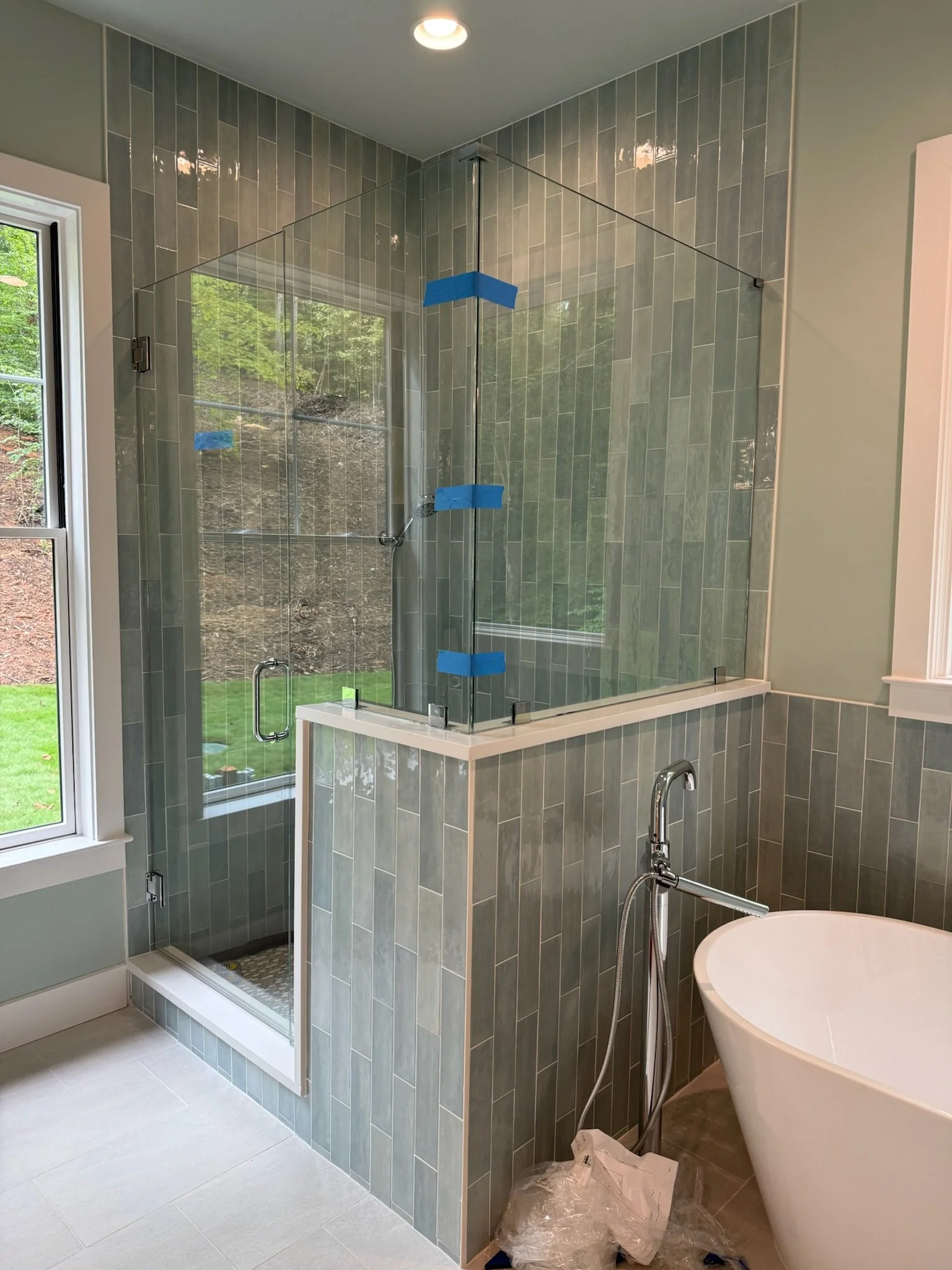 Bathroom with a walk-in shower enclosed by glass panels and a bathtub, with a window showing greenery outside, and blue painter's tape on the shower glass.