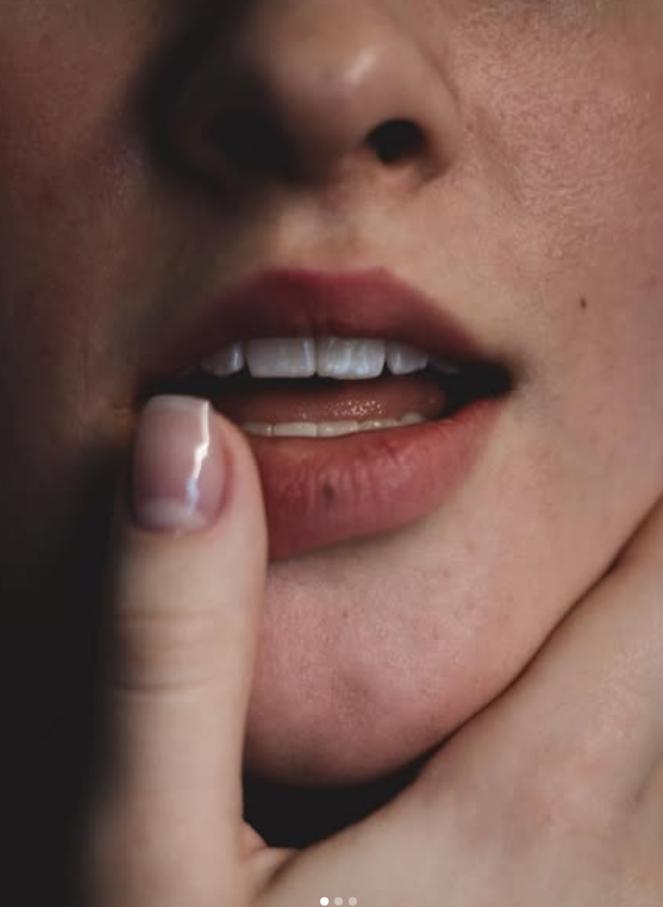 Gros plan sur une bouche féminine légèrement ouverte, avec le doigt posé sur la lèvre inférieure, montrant les dents et la commissure de la bouche.