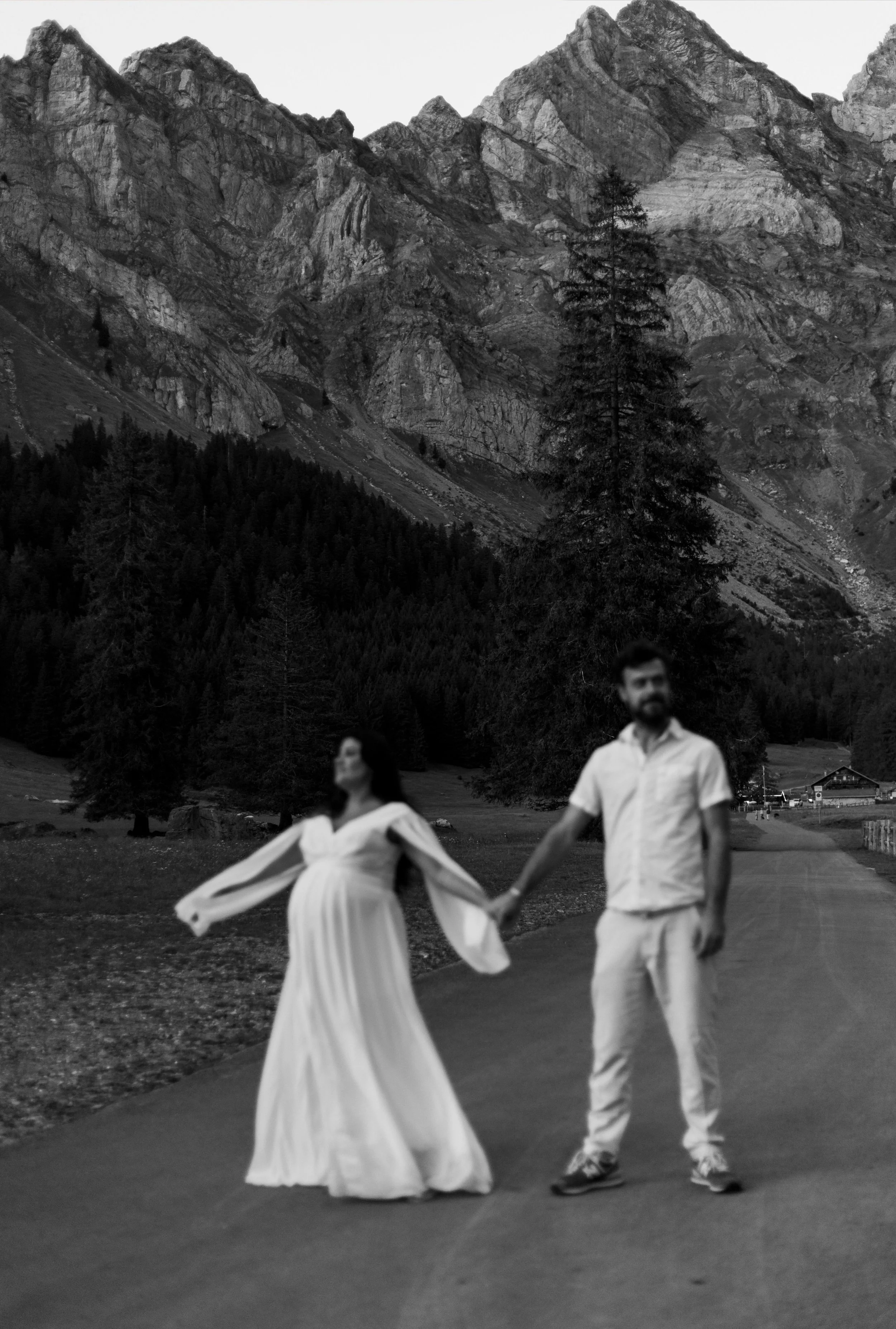 Une photo en noir et blanc d'un couple marchant sur une route dans un paysage de montagne avec des forêts de conifères et des montagnes escarpées en arrière-plan.