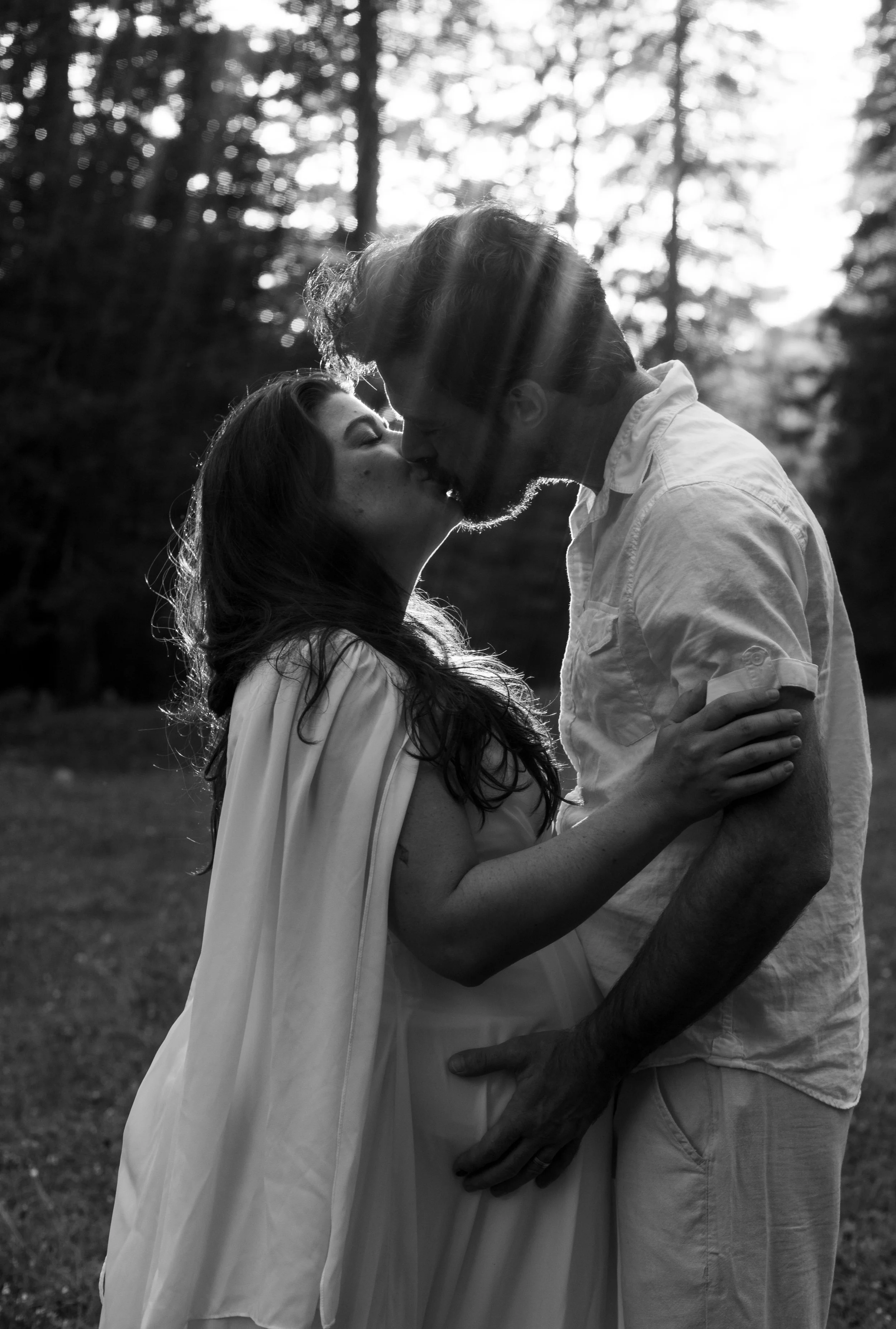 Un couple s'embrasse dans une forêt, en noir et blanc, avec la lumière du soleil qui filtre à travers les arbres.