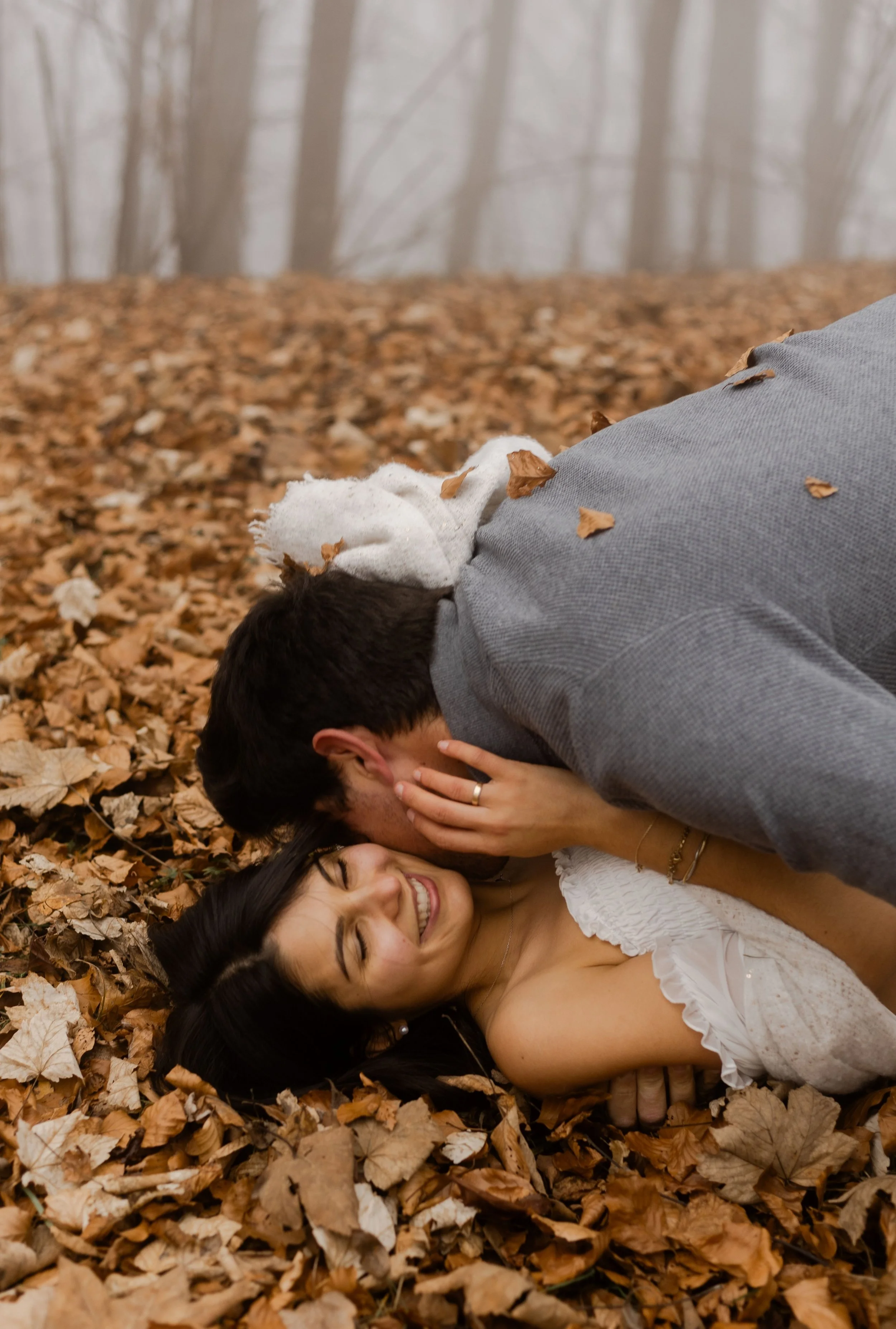 Un couple dans une forêt automnale, la femme allongée sur un tapis de feuilles mortes, souriant tandis que l'homme lui donne un baiser sur le front.