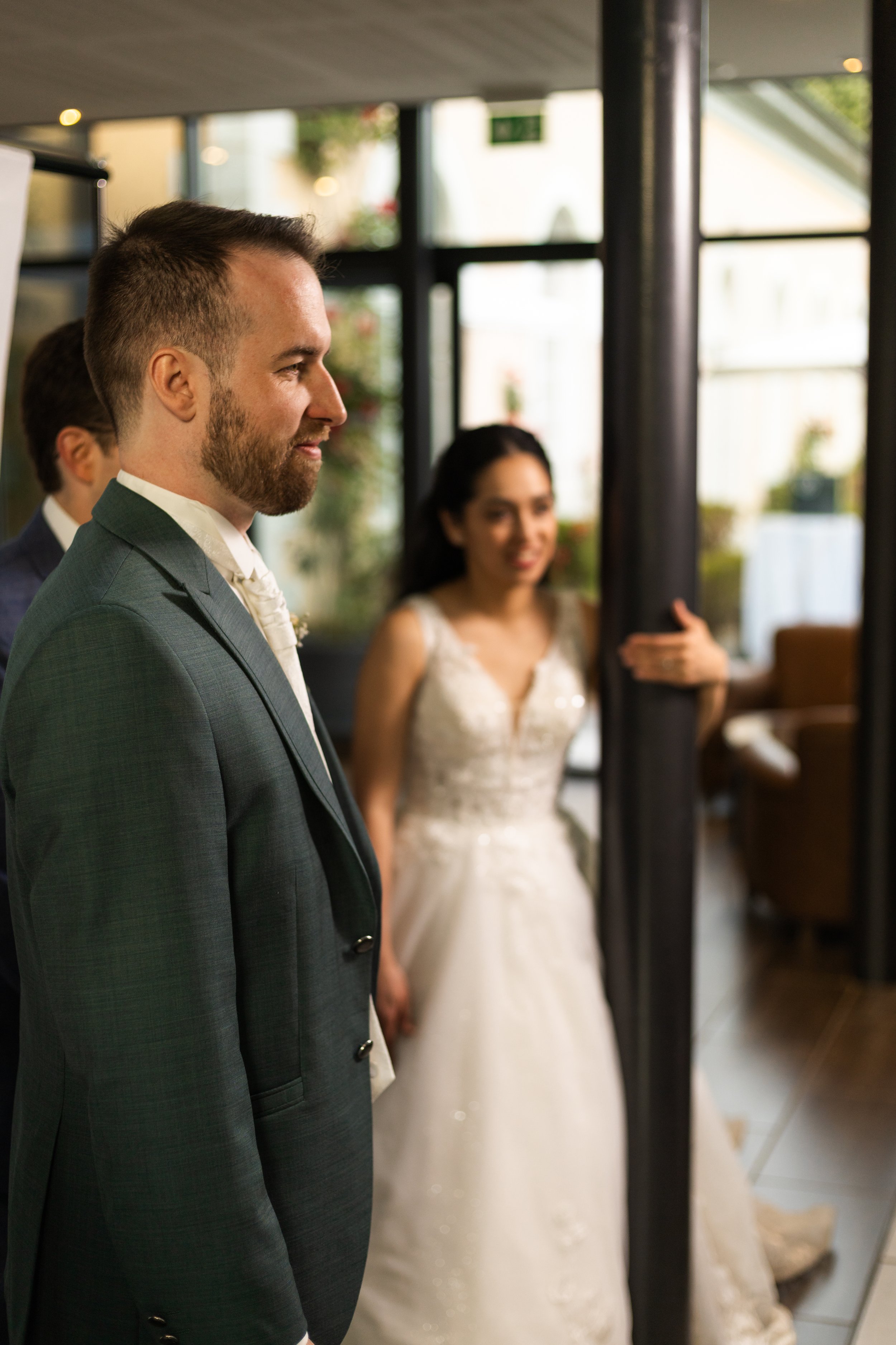 Un homme avec une barbe et une veste en costume gris regarde à gauche, derrière lui, une femme en robe de mariée blanche sourit et fait un geste de la main, dans un décor lumineux avec des fenêtres.