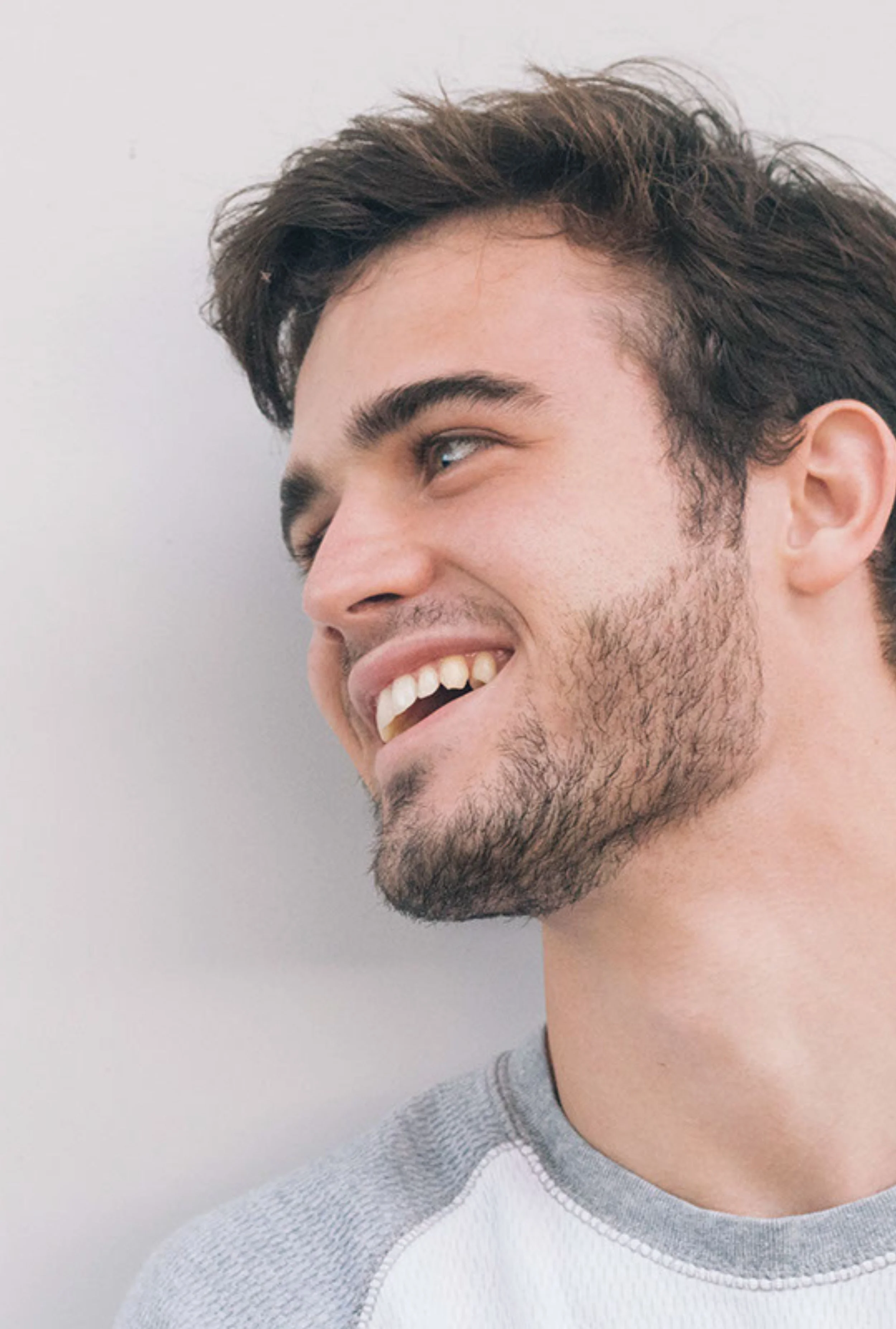 Jeune homme souriant avec barbe, vu de côté, portant un t-shirt gris et blanc.