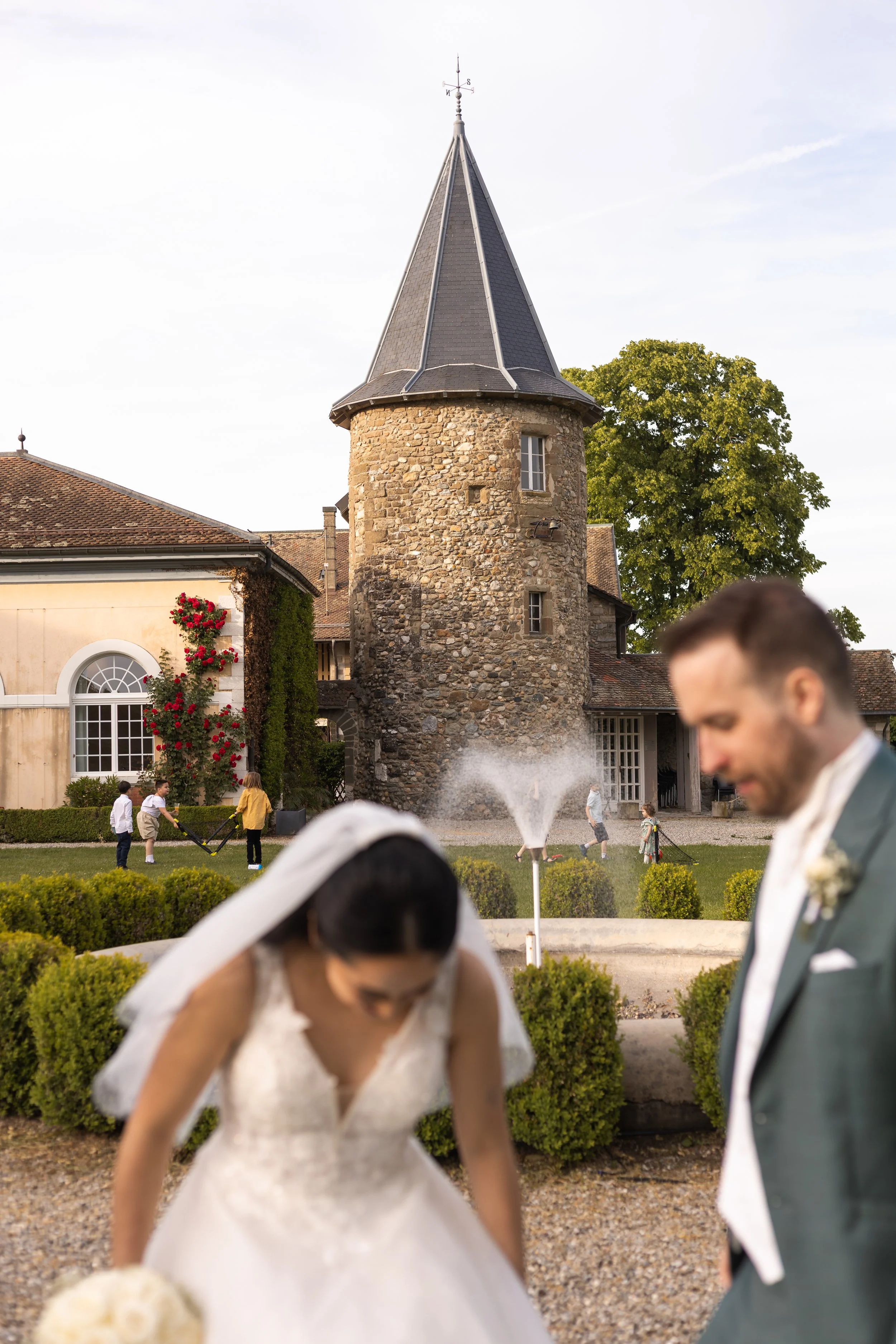 Une cérémonie de mariage à l'extérieur avec un bâtiment en pierre en arrière-plan, des enfants jouant et un couple de mariés au premier plan, la mariée en robe blanche et le marié en costume gris. 