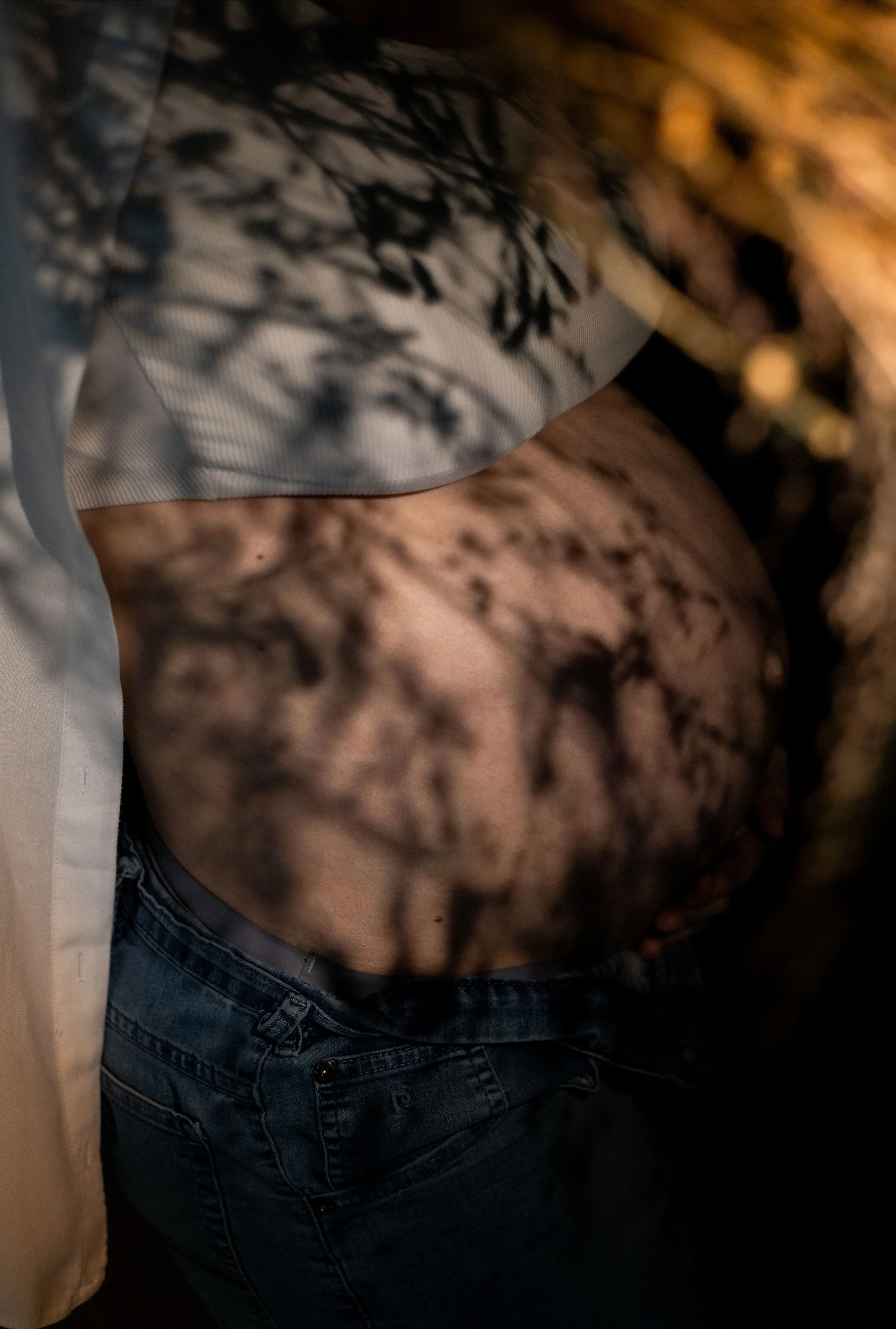 Partie abdominale d'une personne portant un t-shirt à motifs et un jean, avec des ombres de feuilles projetées sur le corps.