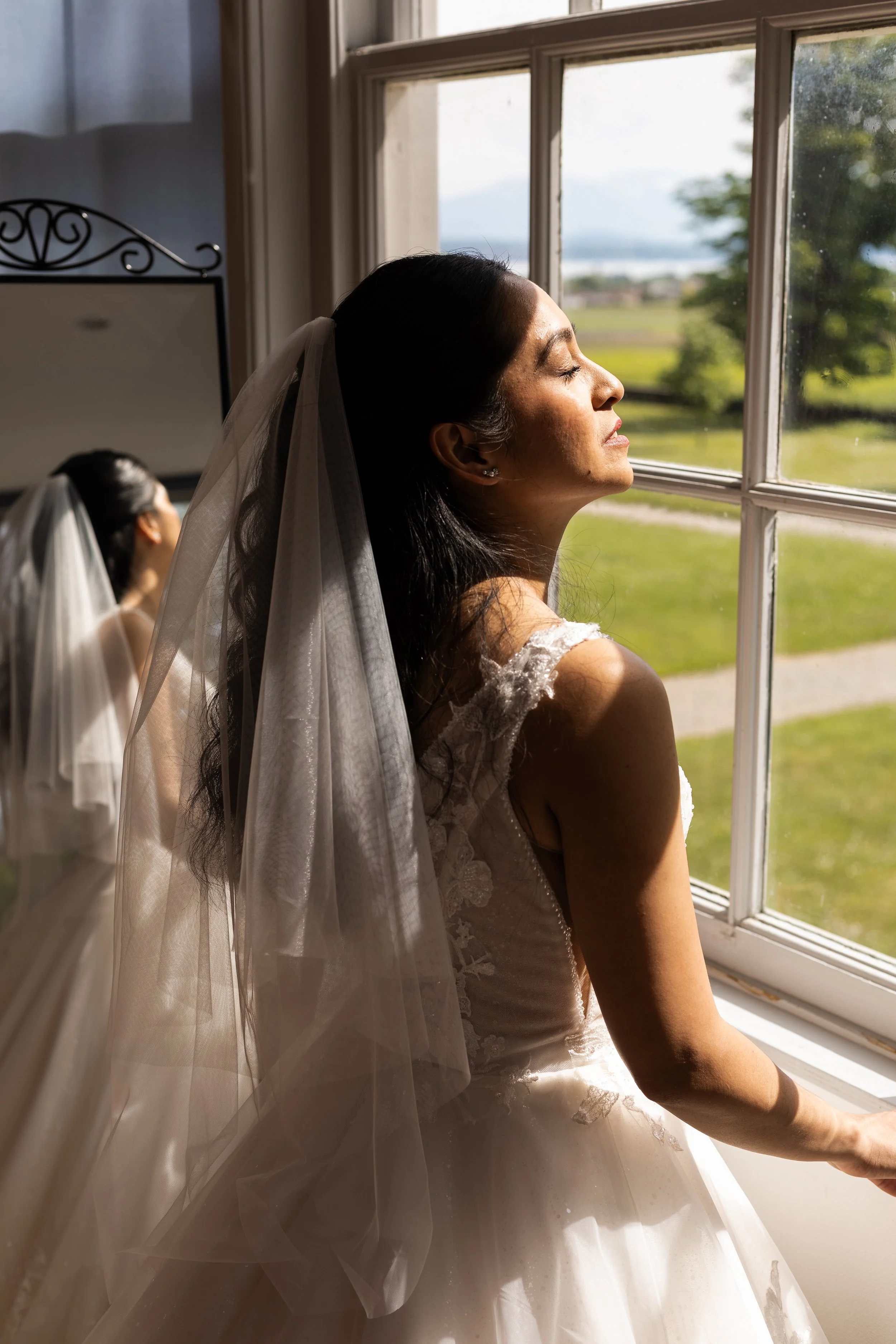 Une femme en robe de mariée regarde par la fenêtre, avec la lumière du soleil illuminant son visage, dans une pièce avec une vue verte à l'extérieur.