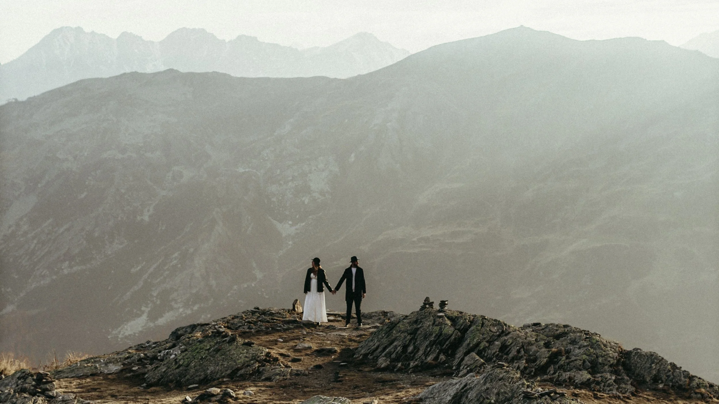 Un couple habillé en noir et blanc, tenant la main, se tient sur une roche en montagne, avec un paysage de montagnes en arrière-plan.