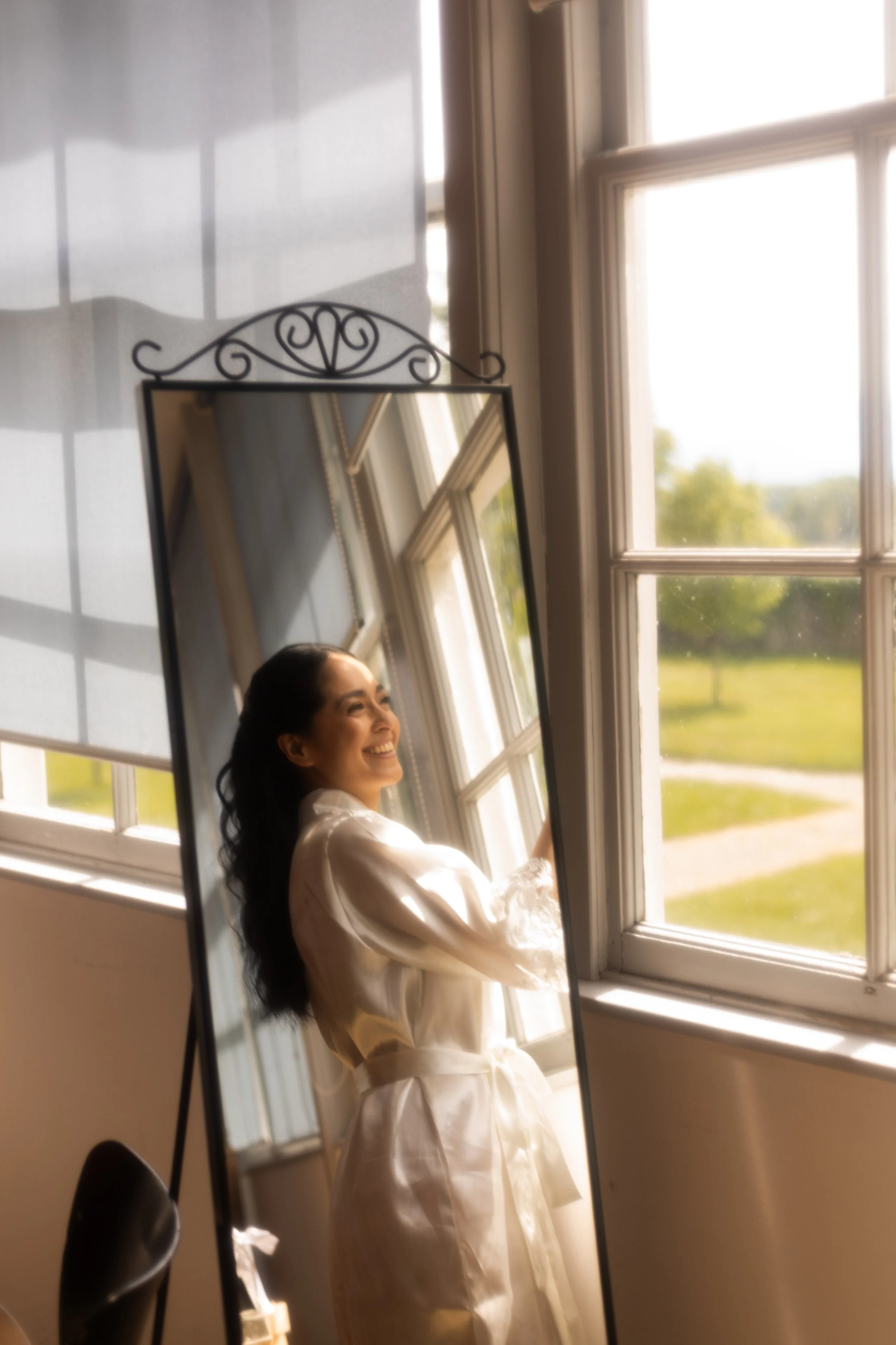 Une jeune femme souriante portant une robe blanche, vue dans un miroir, près d'une grande fenêtre avec un paysage verdoyant en arrière-plan.