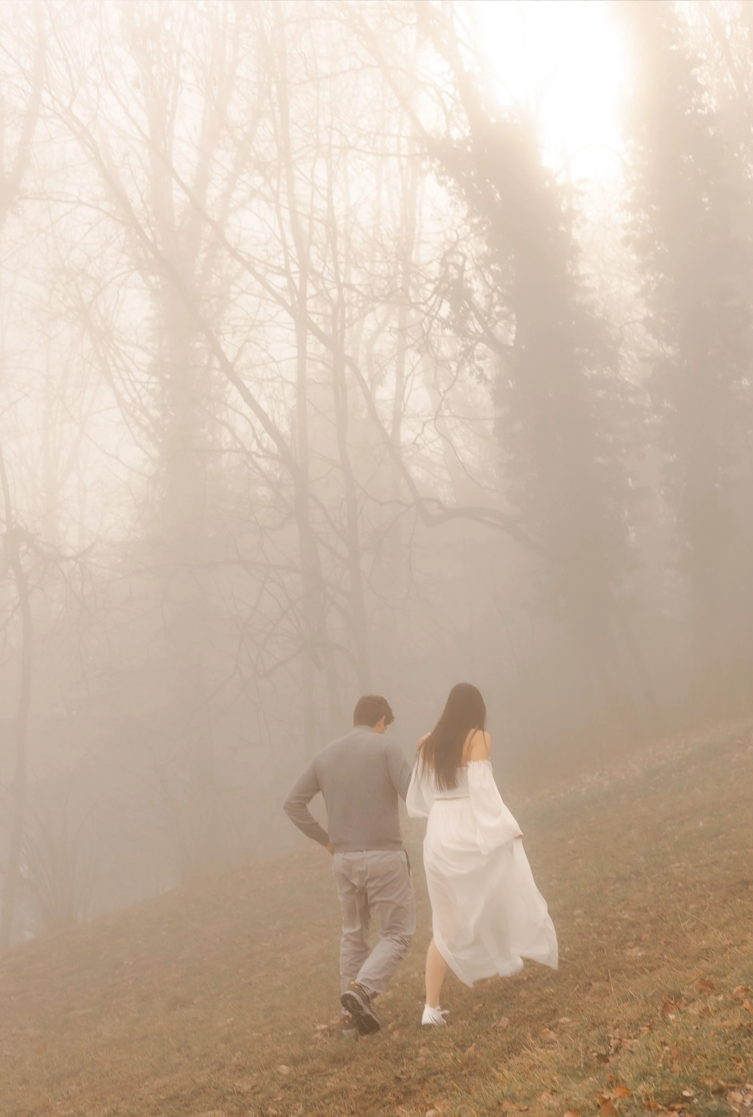 Un homme et une femme marchent dans une forêt brumeuse. La femme porte une robe blanche fluide, et l'homme porte un pull gris et un pantalon clair.
