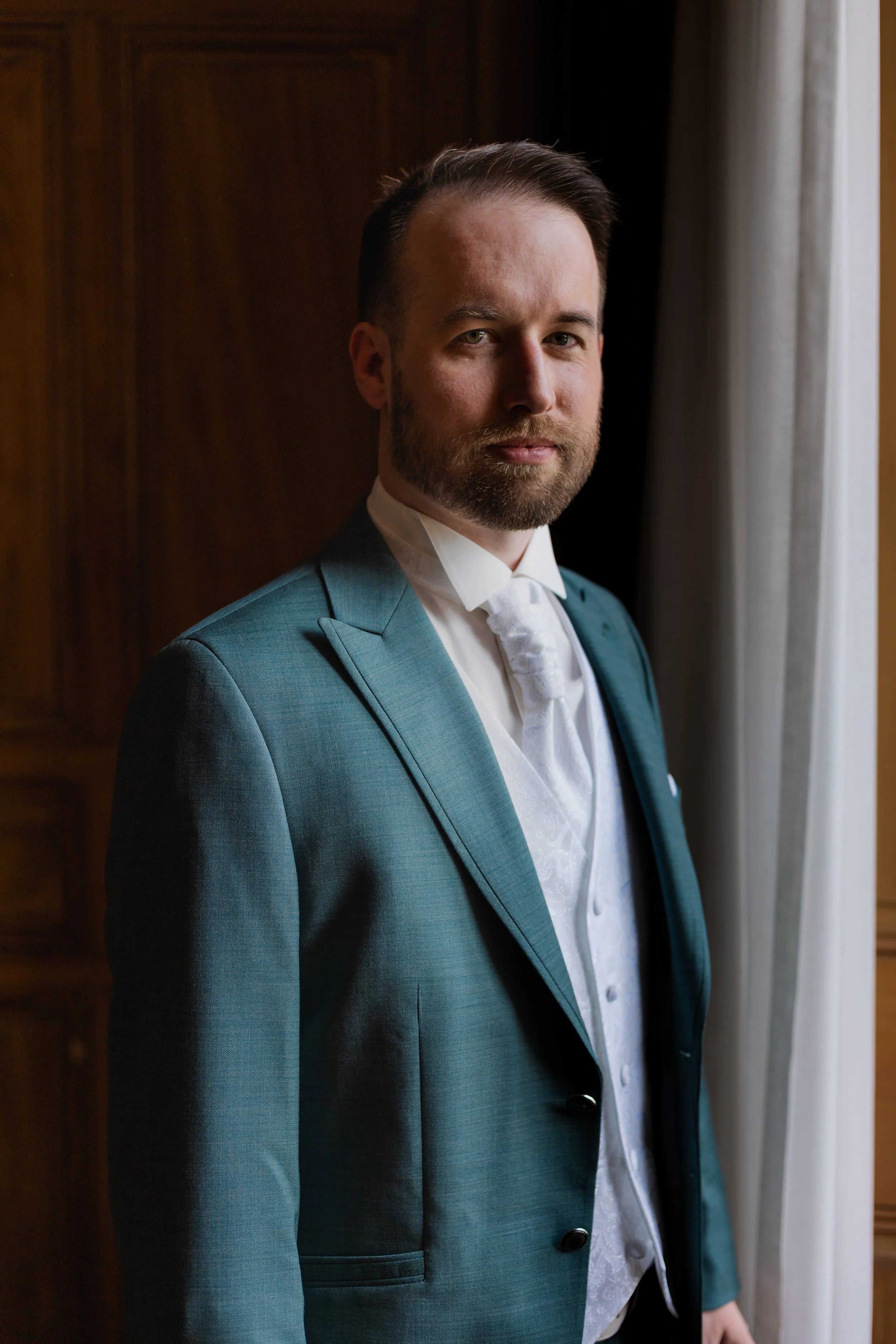 Homme en costume bleu avec chemise blanche, debout près d'une fenêtre avec rideaux, intérieur chaleureux.