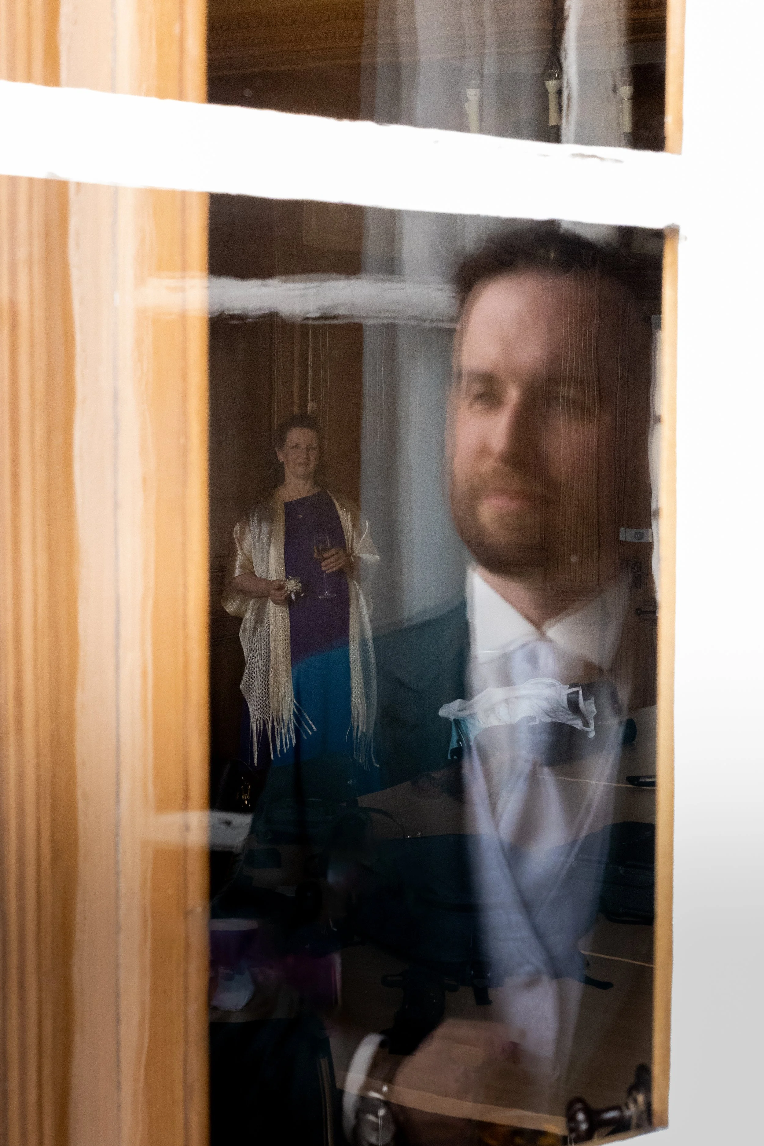 Reflet d'une personne assise devant un miroir, avec une femme tenant un verre de champagne en arrière-plan visible à travers la vitre du miroir.