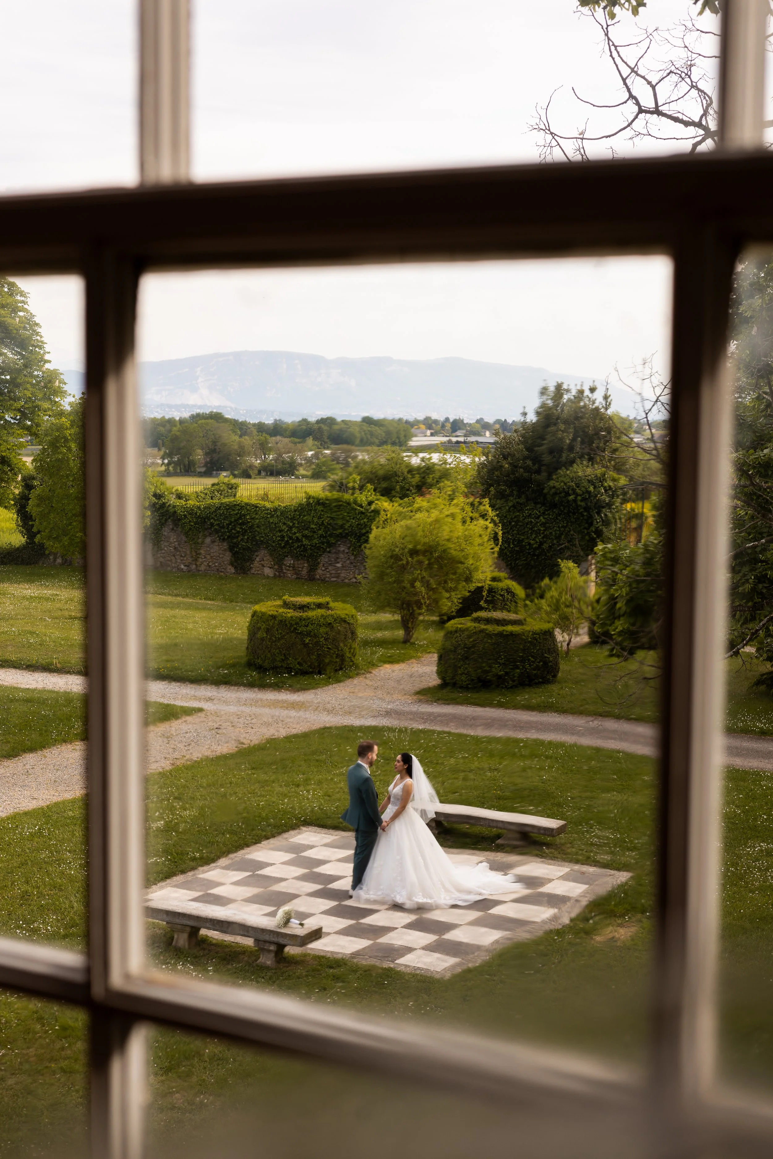 Un couple de mariés se tient par la main sur une terrasse en pierre dans un jardin. La scène est vue à travers une fenêtre avec des cadres en bois. En arrière-plan, il y a des arbres, des buissons bien taillés, une pelouse verte, et un paysage de mon