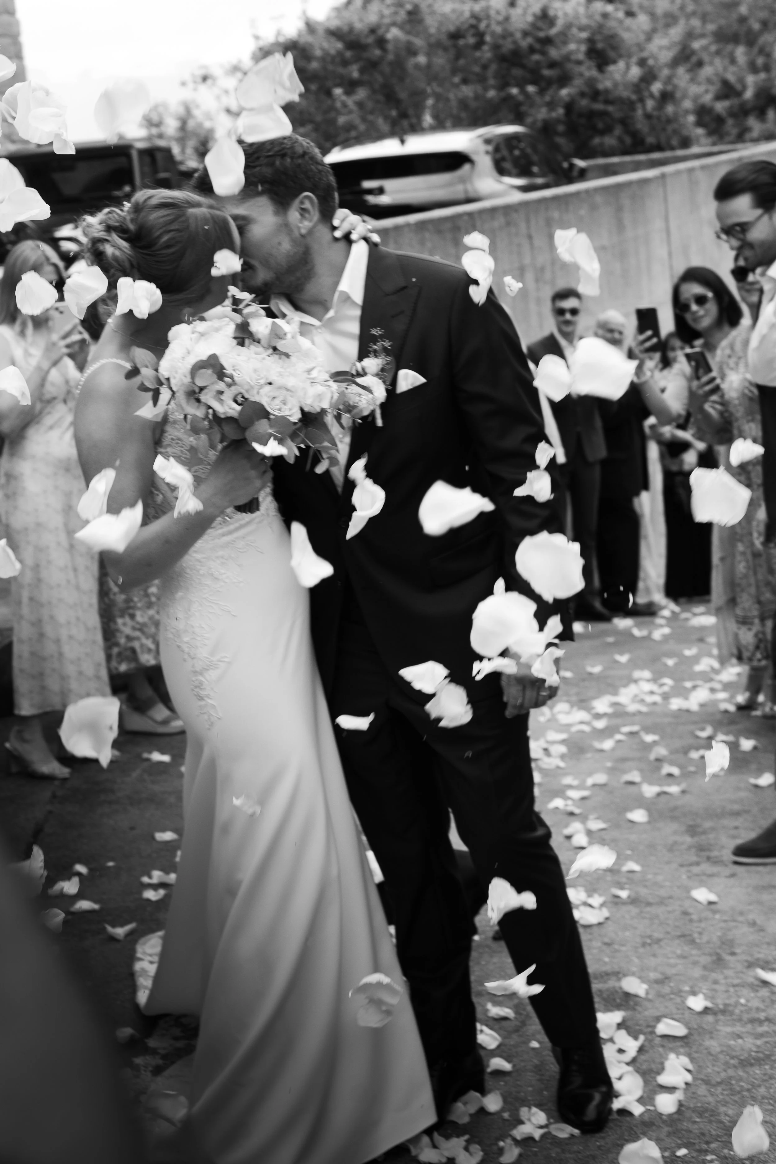 Un couple de mariés s'embrassent lors de leur mariage, entourés d'invités qui jettent des pétales de fleurs, en noir et blanc.
