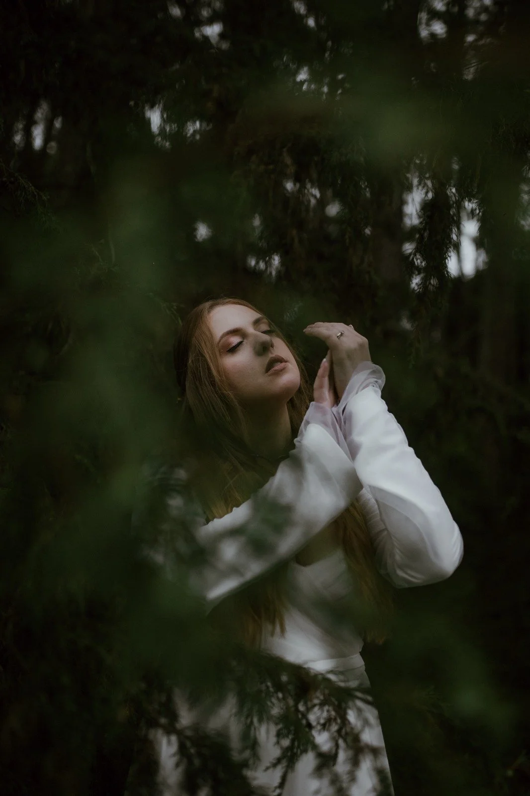 Une femme aux cheveux roux, portant une robe blanche, se tient dans une forêt sombre, avec ses yeux fermés et une expression de contemplation.