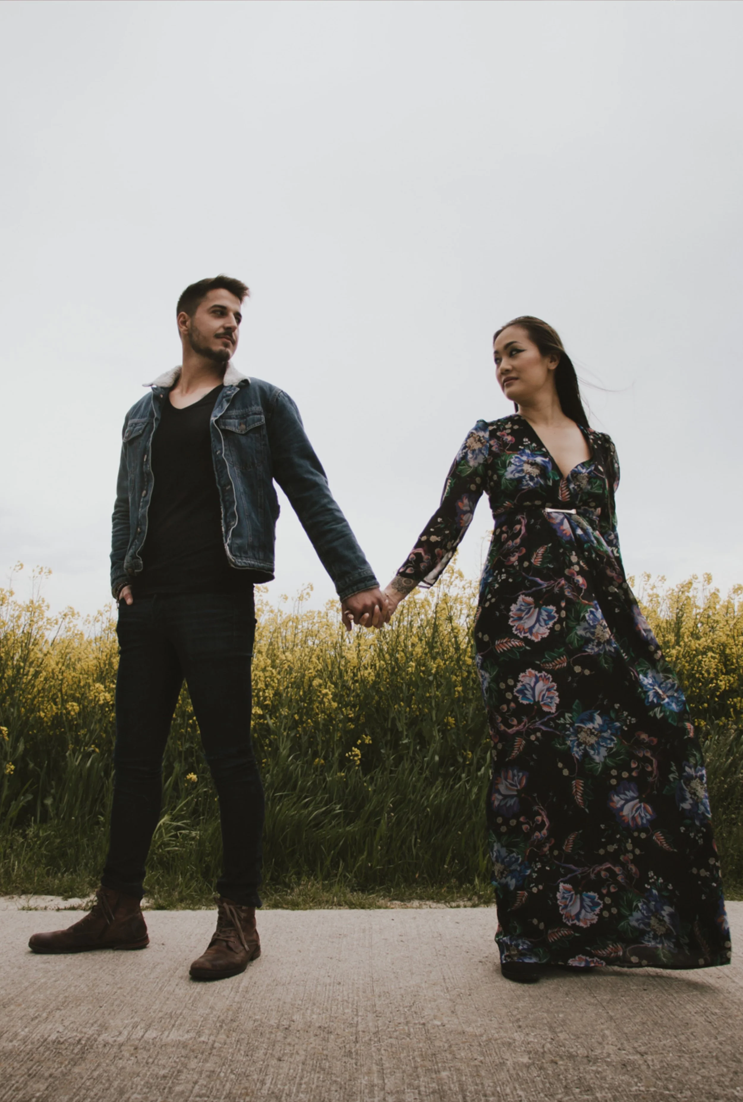 Un homme et une femme se tiennent la main sur un chemin, avec un champ de fleurs jaunes derrière eux, sous un ciel nuageux.