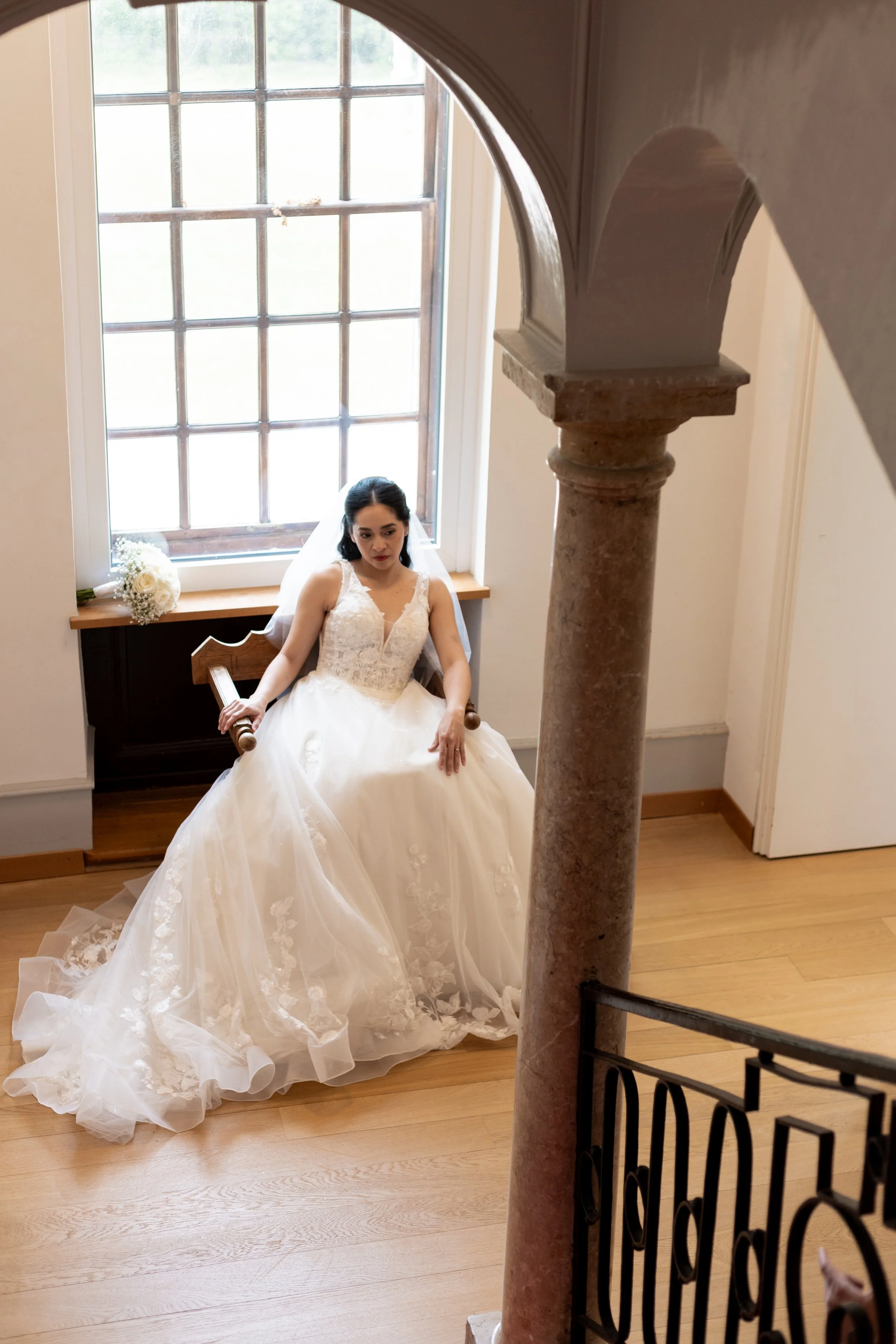 Femme en robe de mariée assise près d'une grande fenêtre avec un bouquet de fleurs blanches à côté, éclairée par la lumière naturelle.