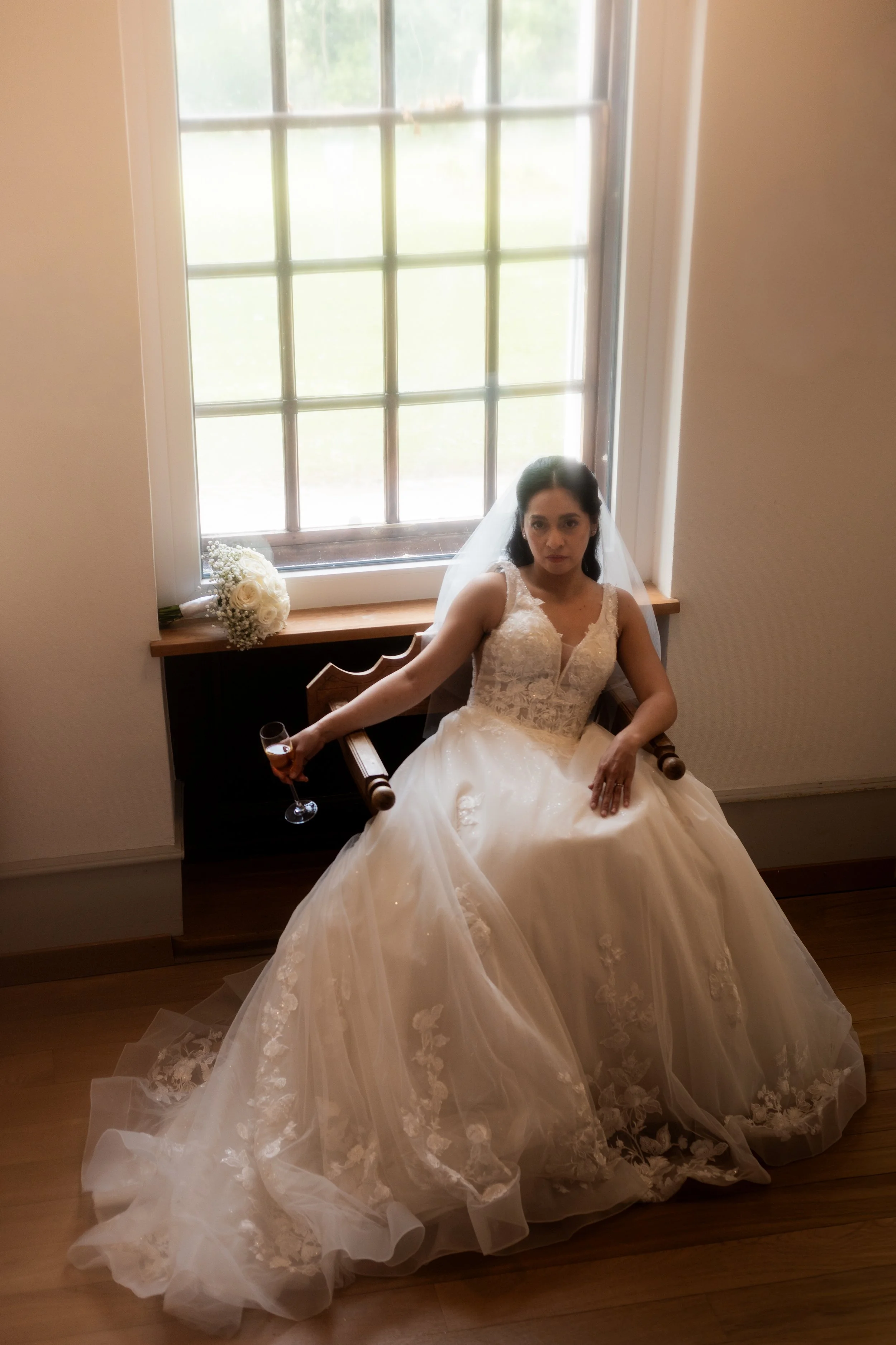 Une femme en robe de mariage assise près d'une grande fenêtre avec des carreaux, tenant un verre de champagne, avec un bouquet de fleurs blanc sur le rebord.